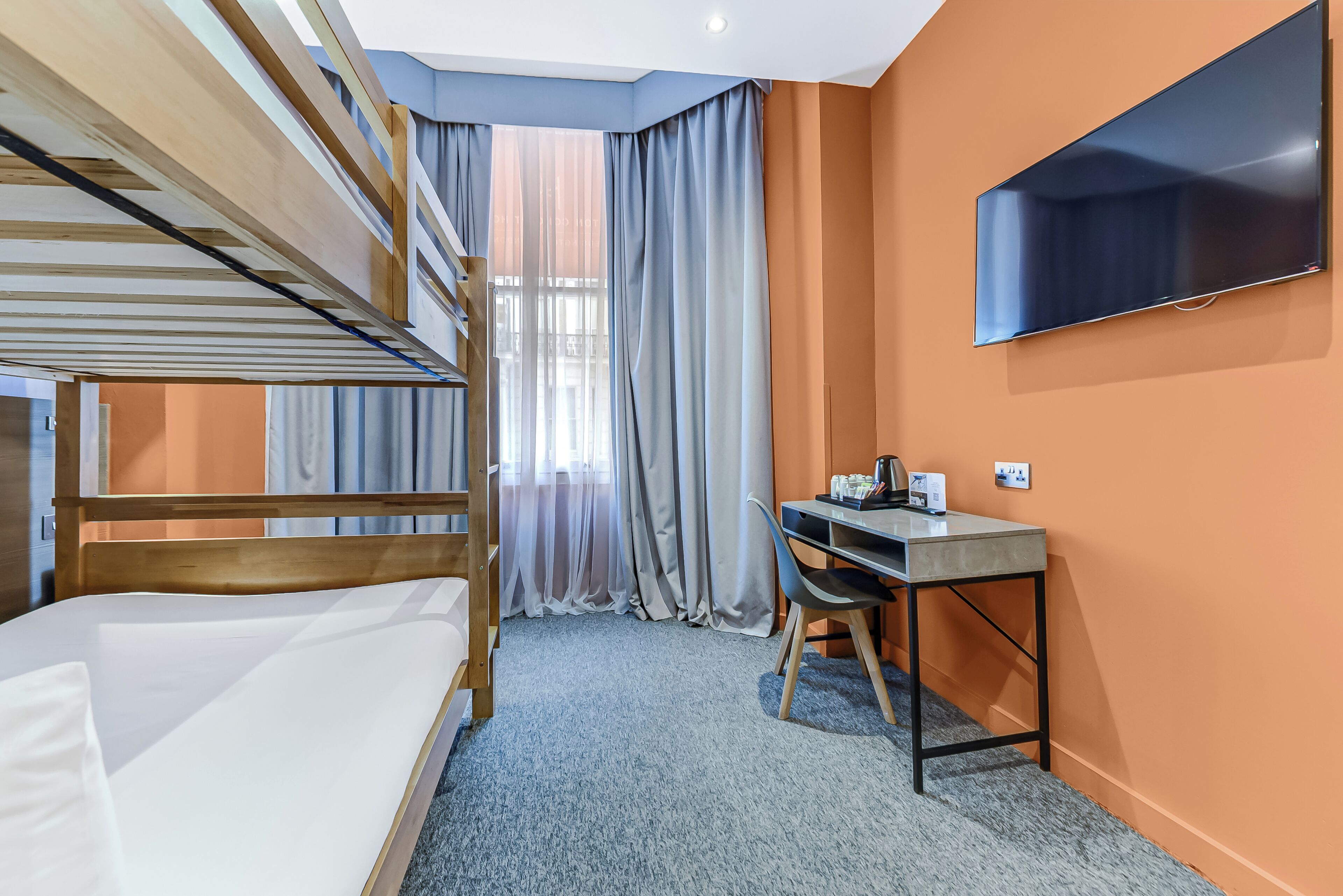 A hotel room with bunk beds, a desk, a chair, a TV, and a window with curtains.