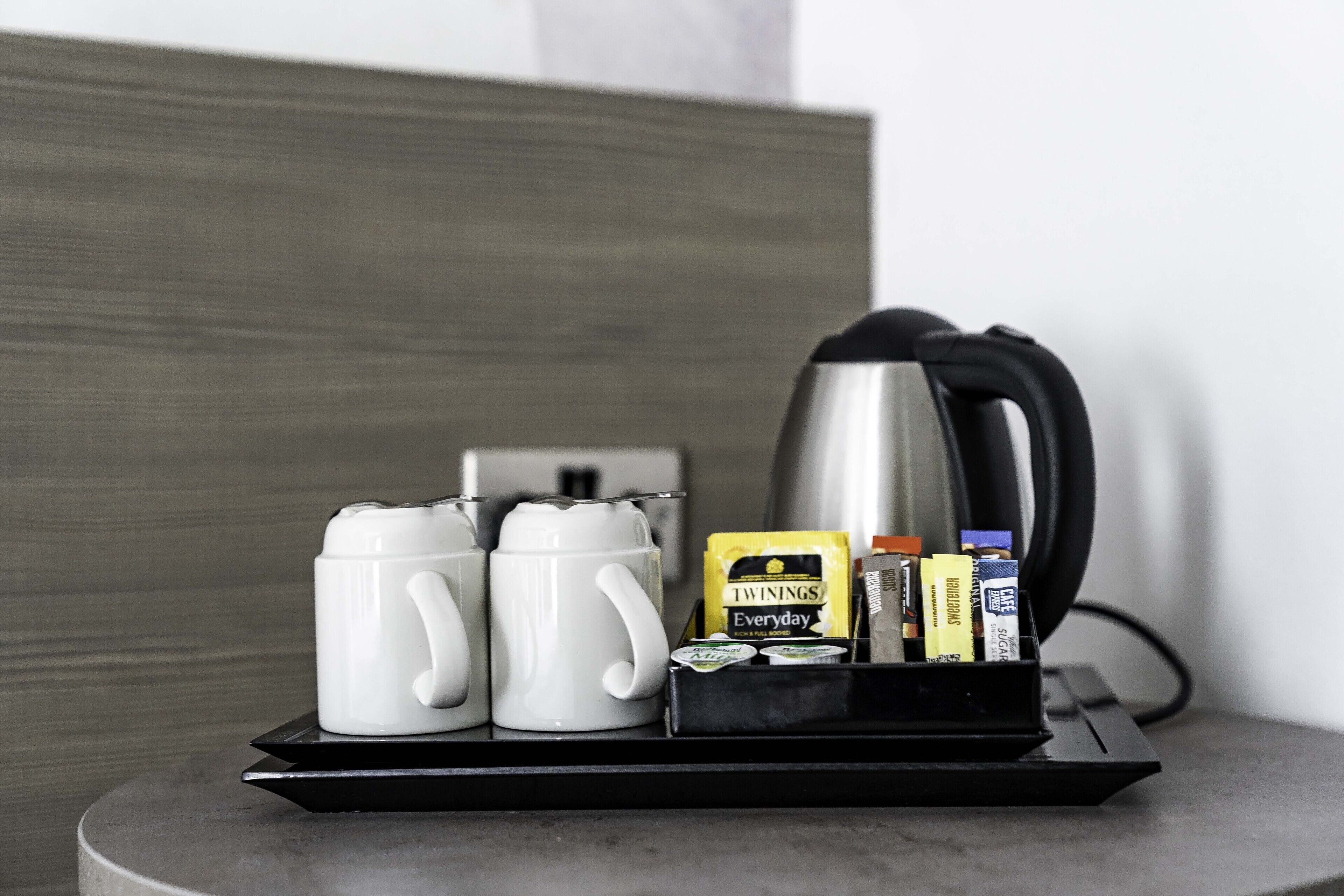 Una bandeja con dos tazas blancas, una cafetera negra, una caja de té Twinings y un portacápsulas de café.