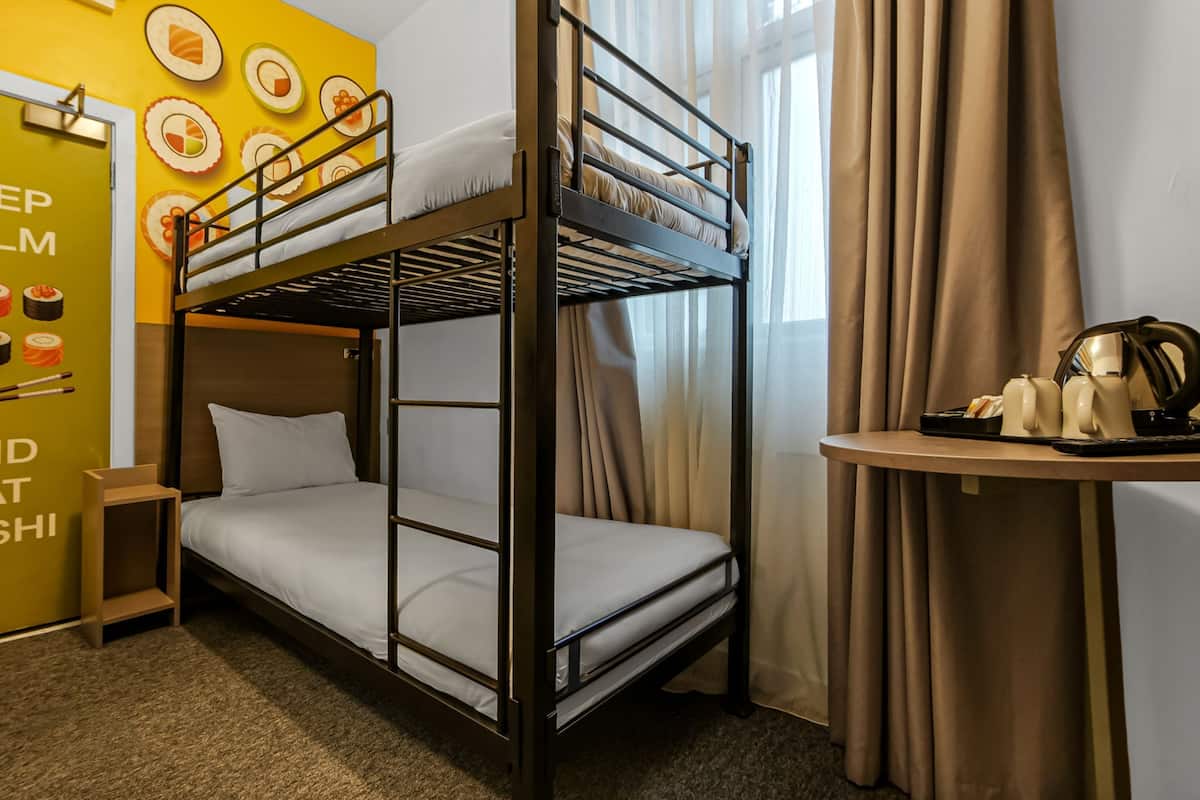 Een slaapkamer met stapelbed, een gele wand met illustraties van sushi en tekst, een klein tafeltje met een waterkoker en een raam met gordijnen.