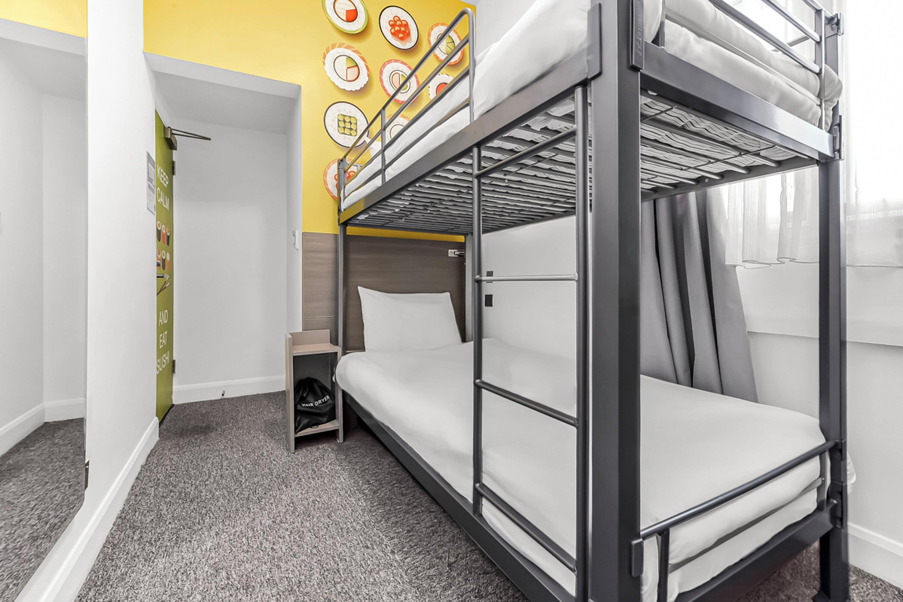 A bunk bed room with a grey carpet, a small wooden shelf, and a wall decorated with food-themed plates.