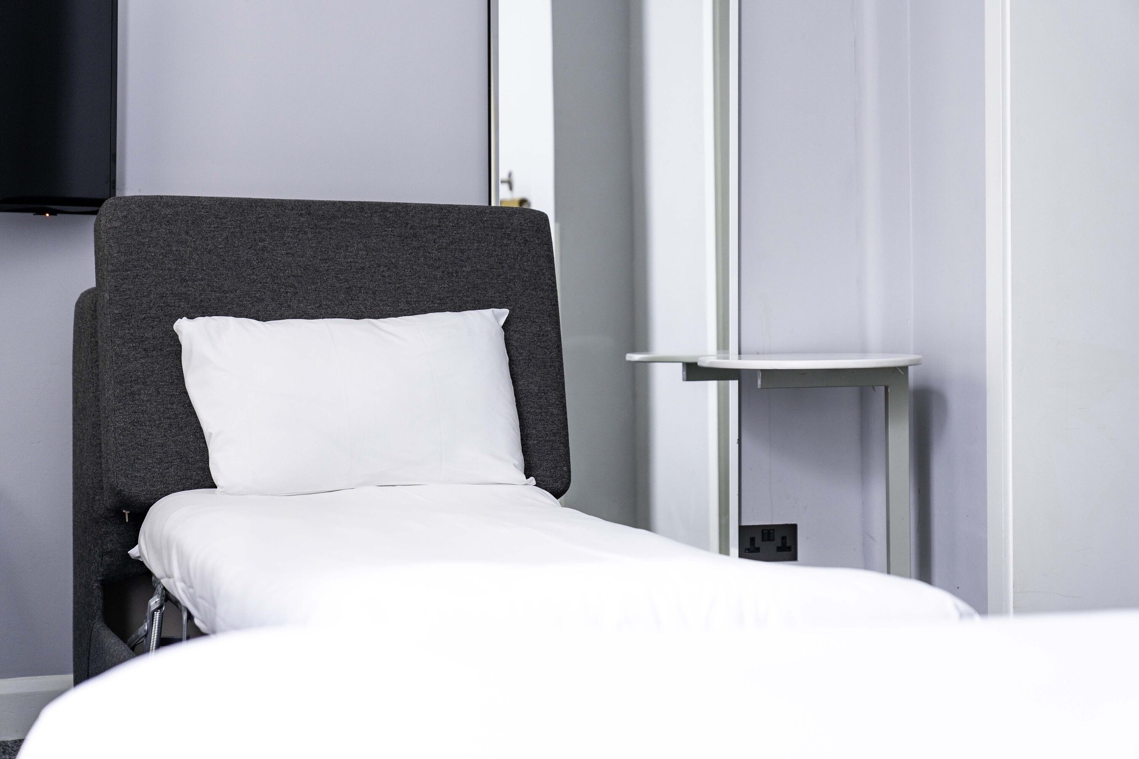 A modern hotel room with a grey upholstered headboard, white bedding, and a small bedside table.