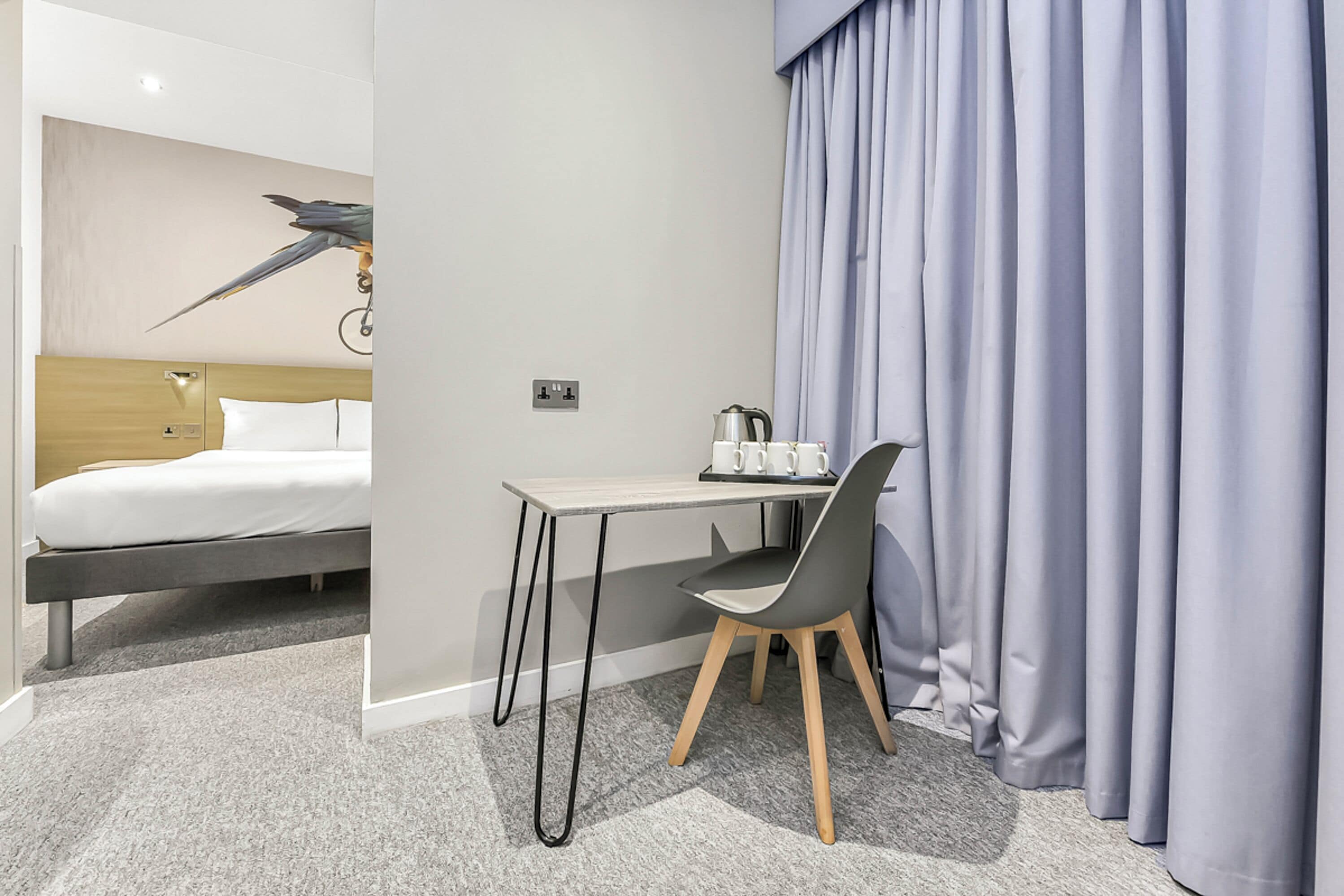 A hotel room with a bed, a desk with a chair, a coffee maker, and a curtain.