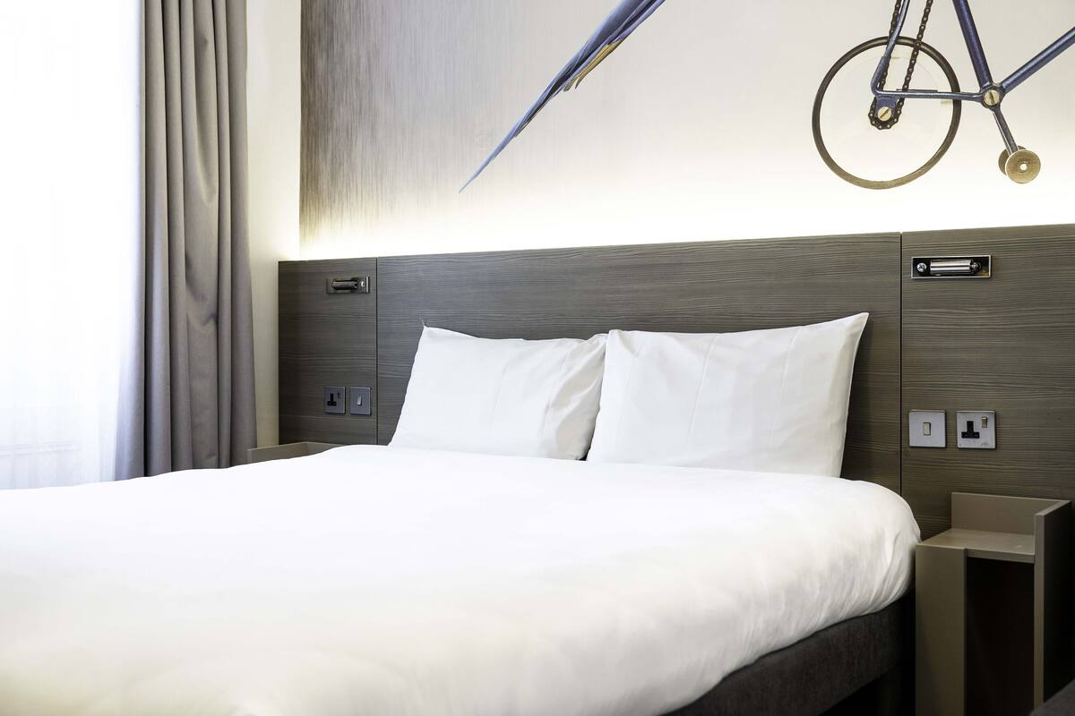 Een moderne hotelkamer met een groot bed, een hoofdeinde met ingebouwde bediening en een aan de muur bevestigde fiets.