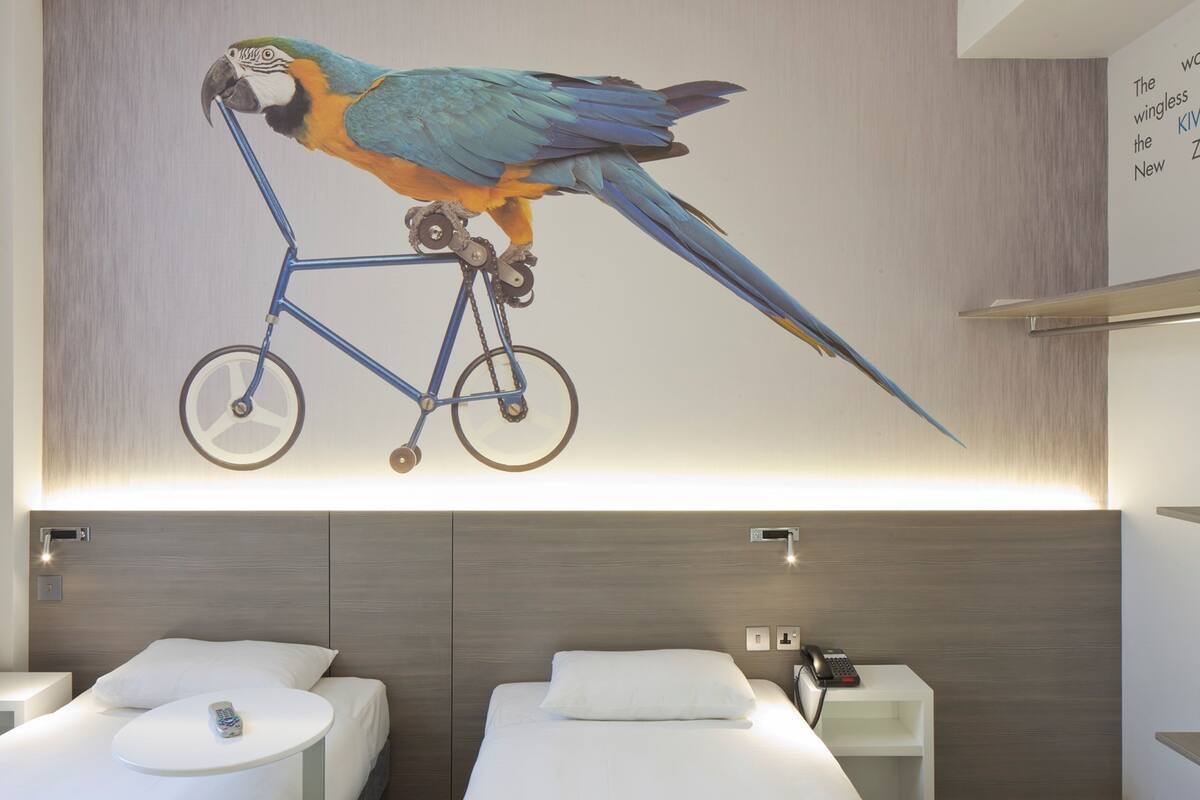 Een moderne hotelkamer met unieke wanddecoratie van een papegaai op een fiets, twee enkele bedden, nachtkastjes en een telefoon op een van de nachtkastjes.