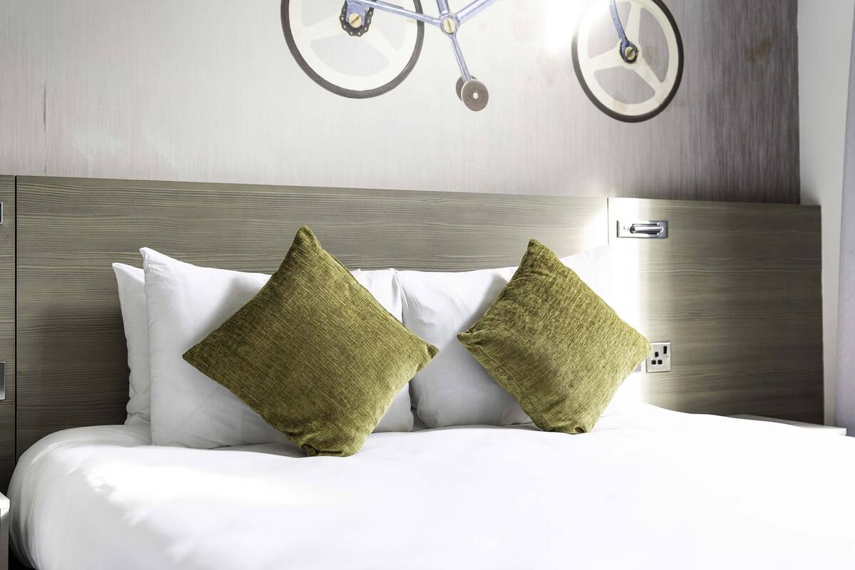 Een net opgemaakt bed met witte lakens en twee groene kussens, een houten hoofdbord en een fietsdecoratie aan de muur.