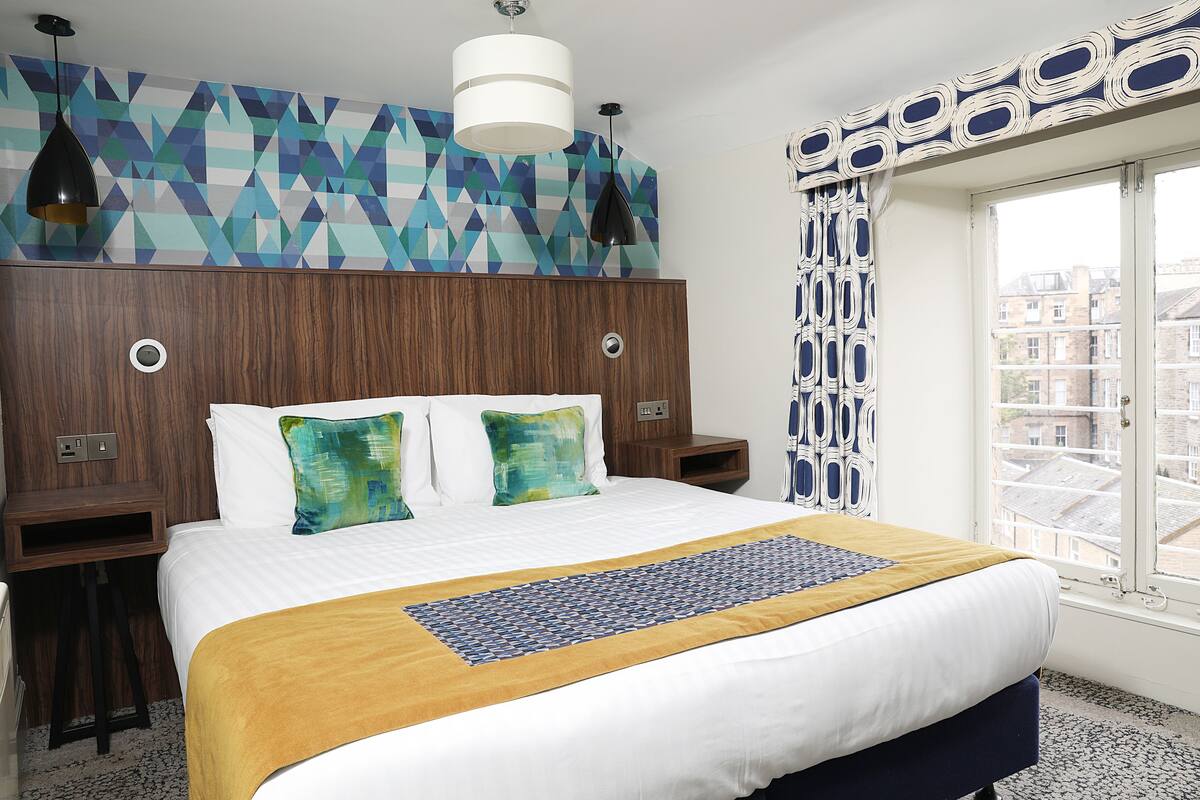 Ett hotellrum med en stor säng, en sänggavel i trä, en vägg med mönster och ett fönster med utsikt över byggnader.