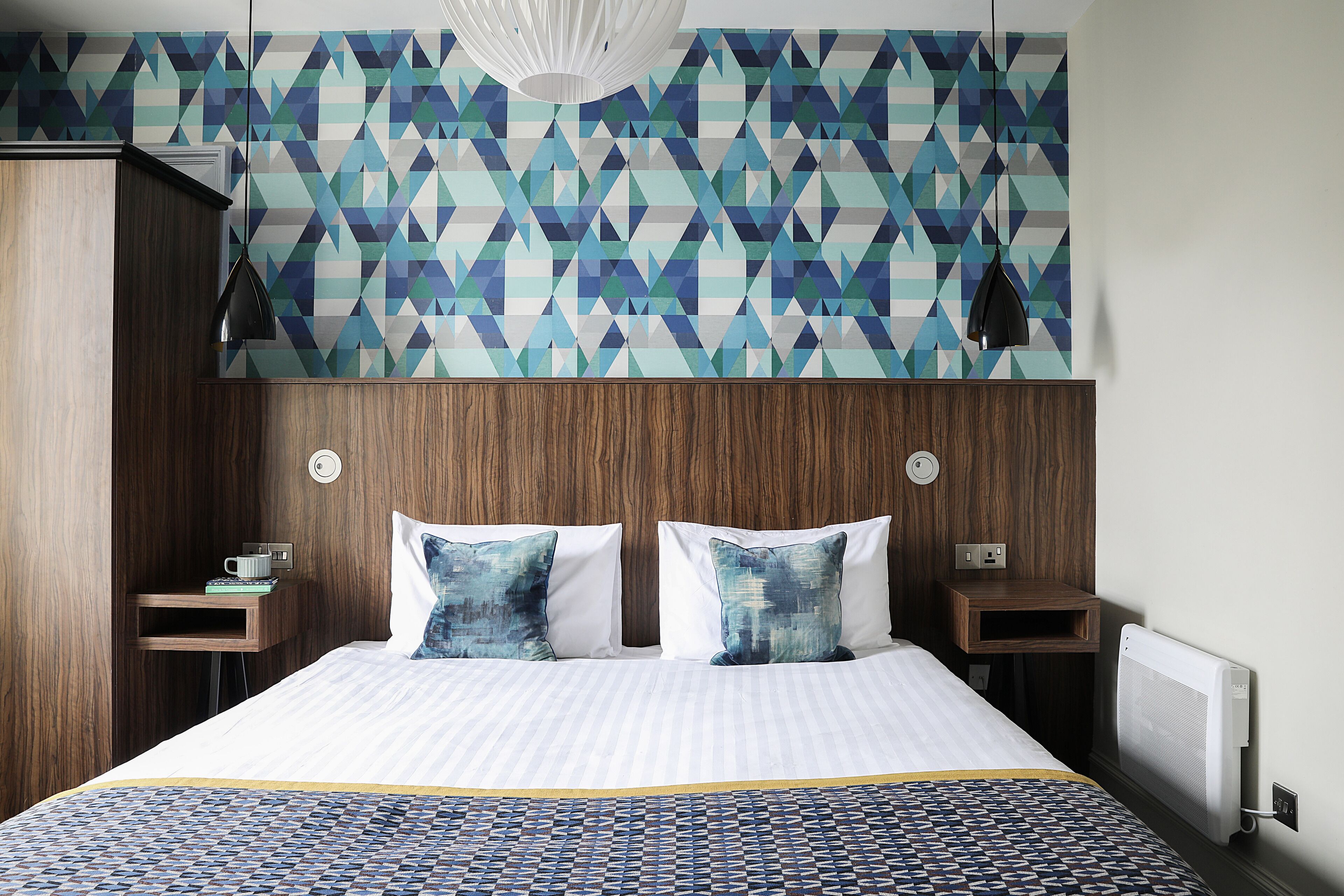 Habitación de hotel moderna con pared de diseño geométrico, cabecera de madera, cama grande con ropa de cama estampada, dos mesitas de noche con lámparas y un radiador fijado a la pared.