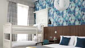 Une chambre avec un lit superposé, un grand lit, un mur à motifs, une fenêtre avec des rideaux et une table de chevet avec une lampe.