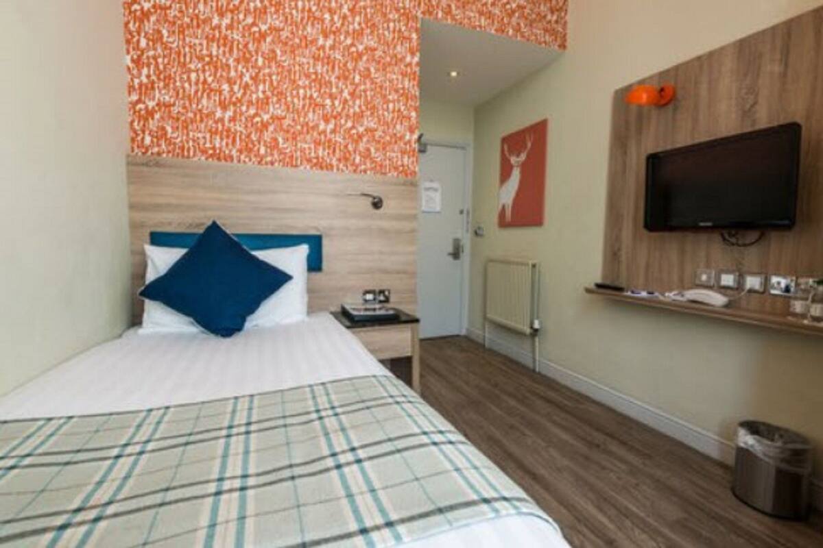 Ett hotellrum med en säng, en tv som är monterad på väggen, ett litet bord och en papperskorg.