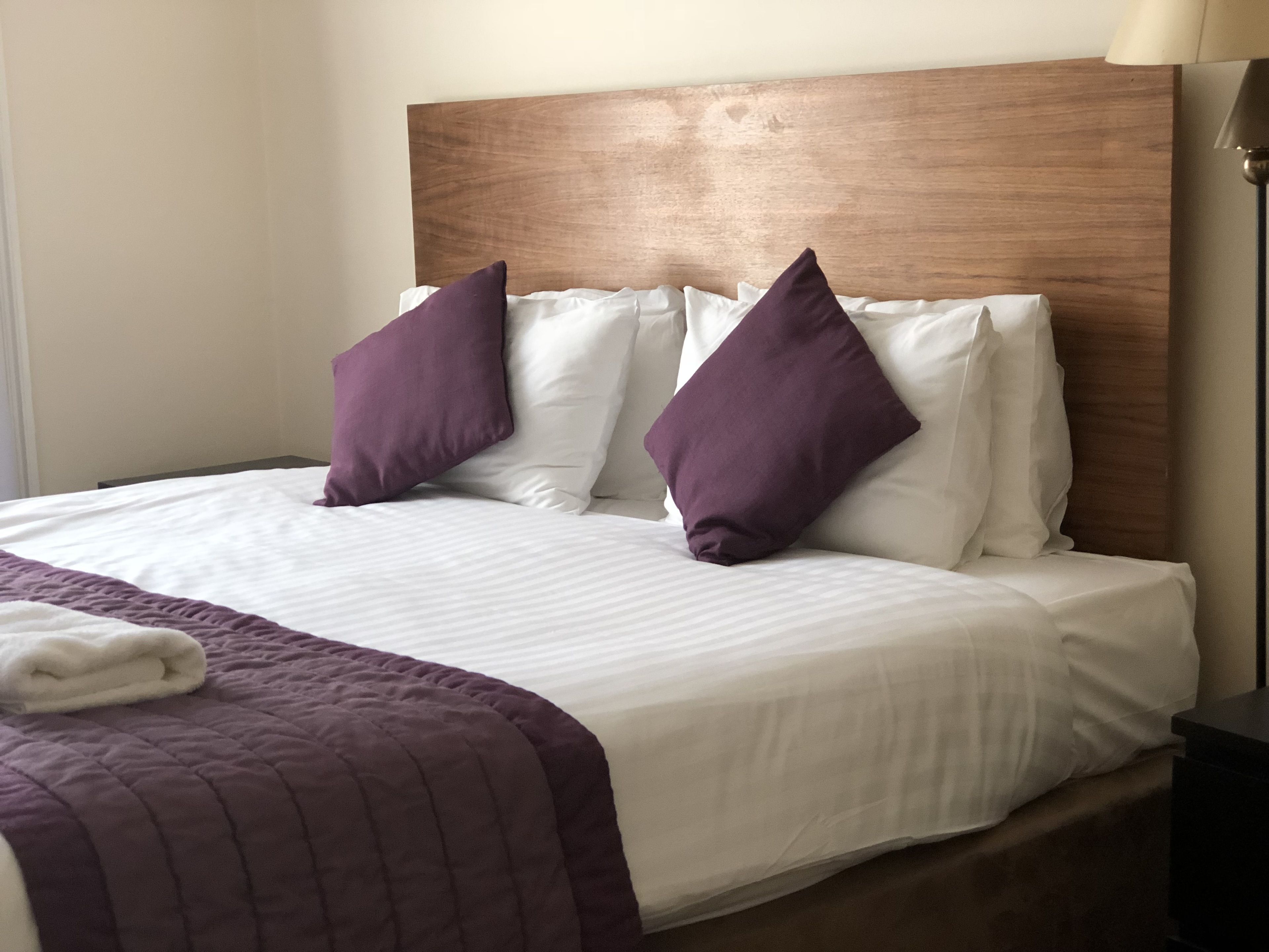 A neatly made bed with white and purple bedding, a wooden headboard, and a lamp beside it.