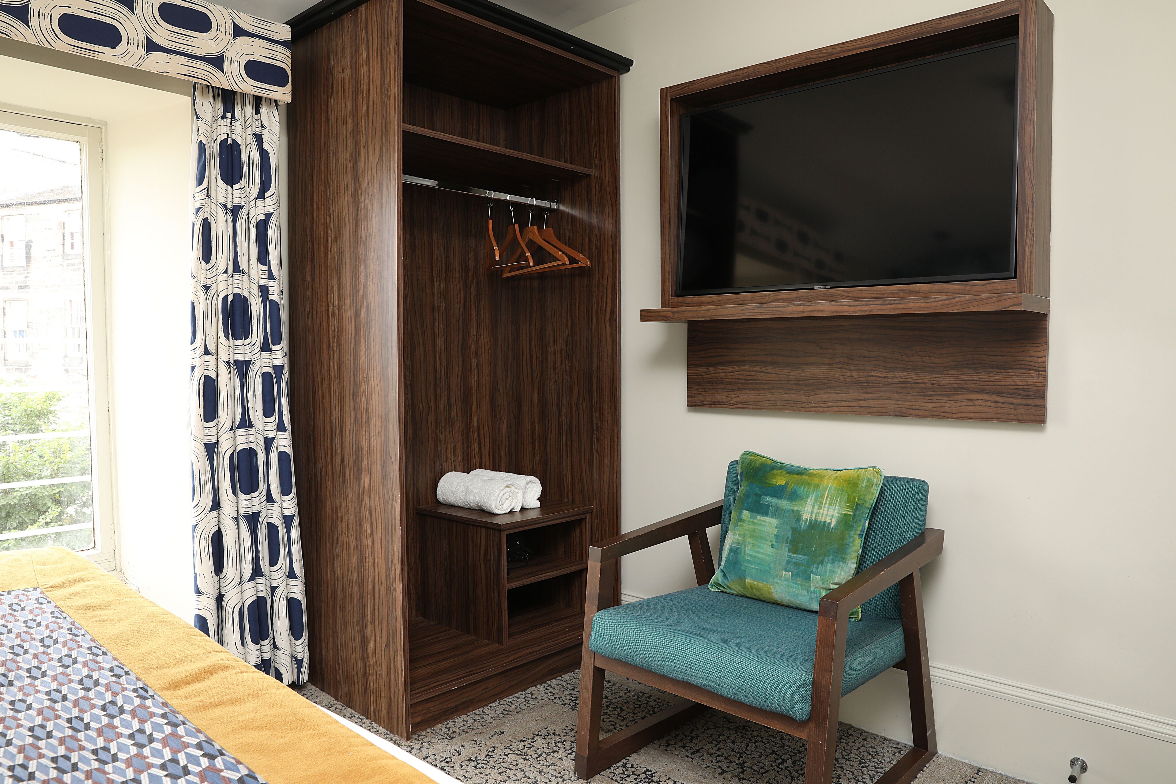 Una habitación con una cama, un armario de madera, una silla con cojín, un televisor montado en la pared y una cortina estampada.