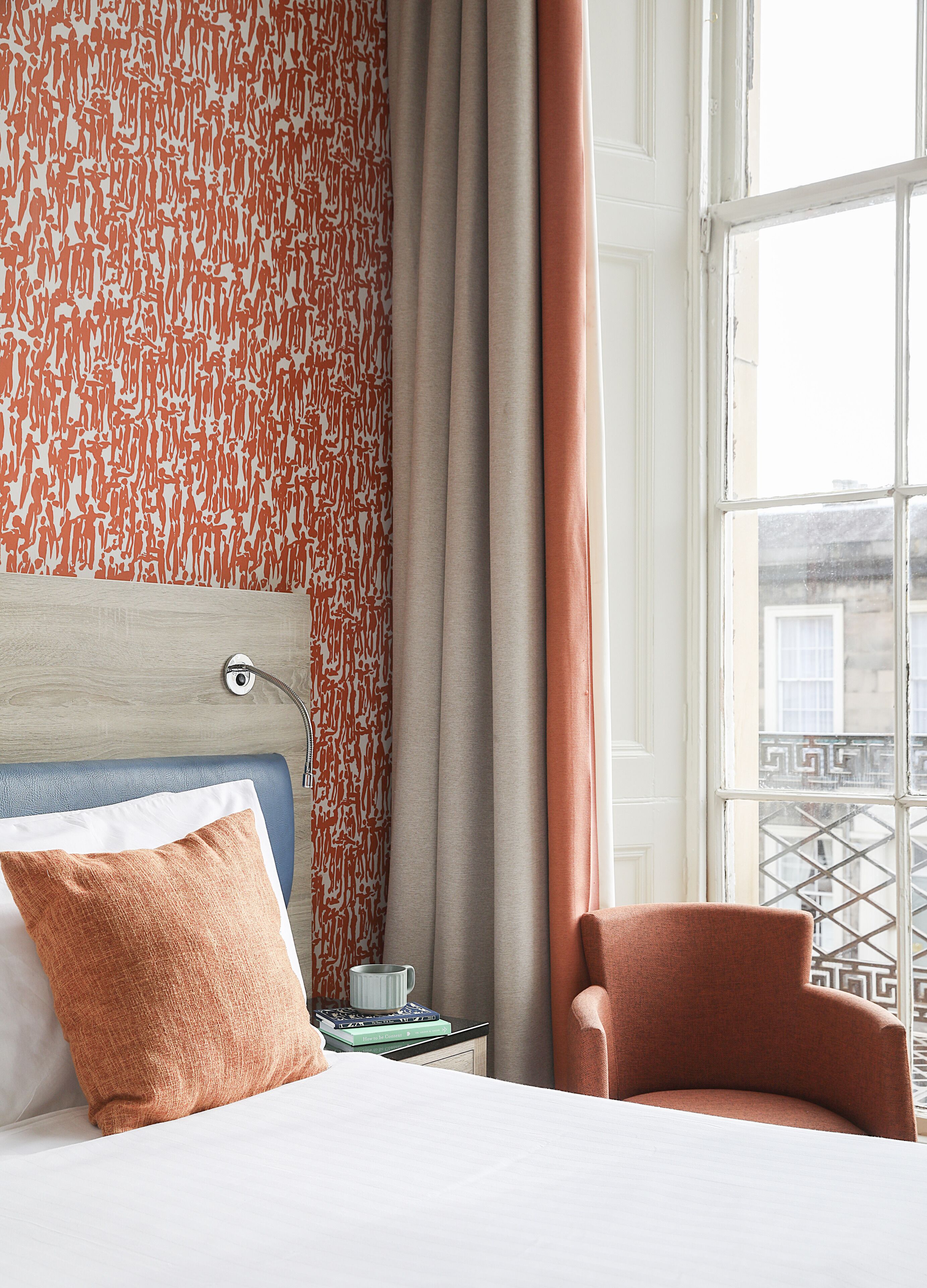 Una habitación de hotel con una cama, una silla, una mesita de noche con libros y una ventana con vistas a edificios.