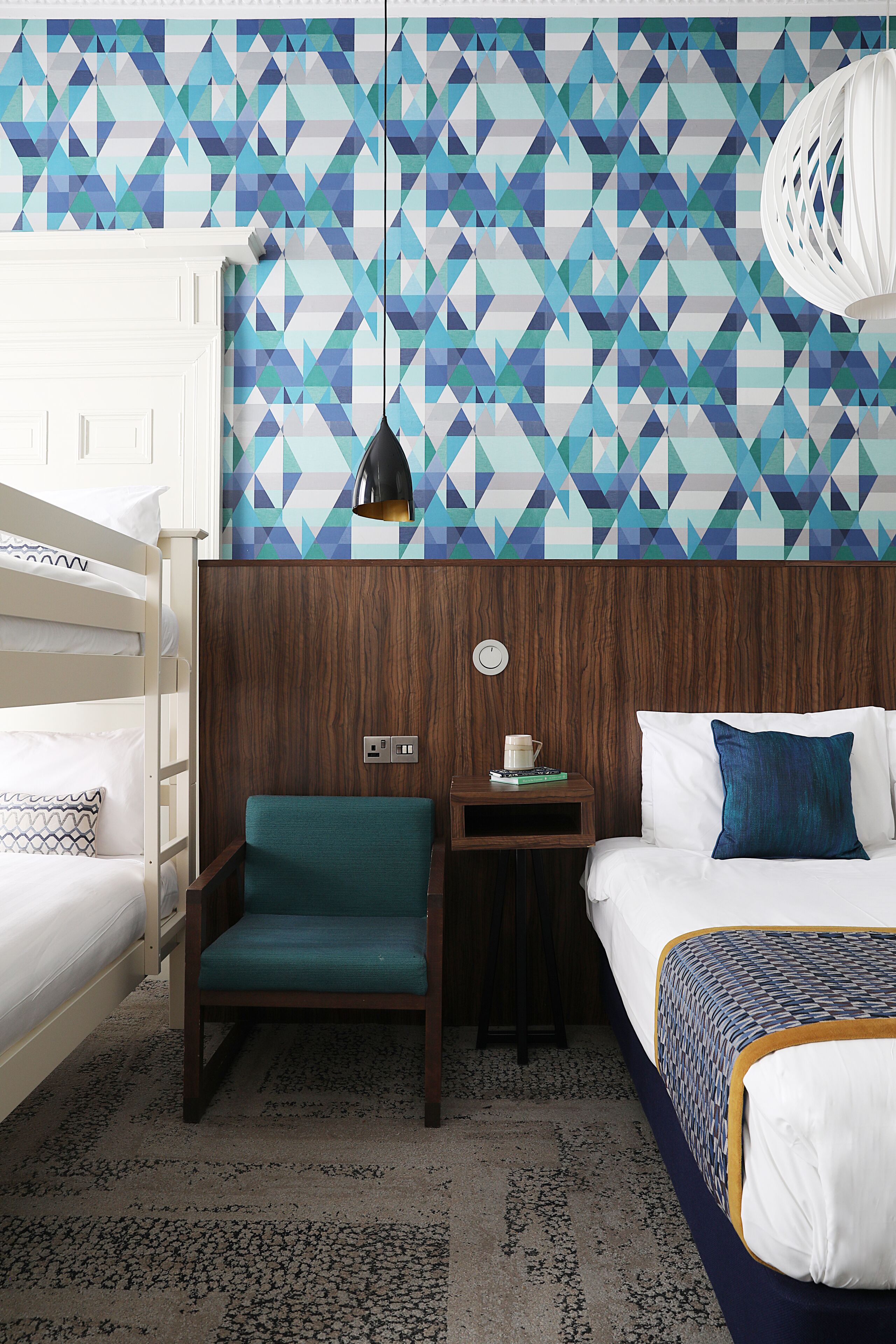 A hotel room with a bunk bed, a single bed, a wooden headboard, a small table with a cup and a book, and a patterned wall.