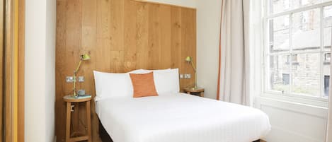Un dormitorio con cabecera de madera, una cama grande con ropa blanca, dos mesitas de noche con lámparas y una ventana con vistas a edificios.