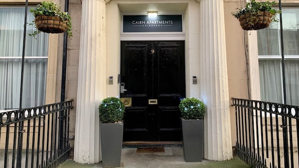 Une entrée d’immeuble avec une porte noire, deux plantes en pot et une enseigne indiquant « CAIRN APARTMENTS EDINBURGH ».