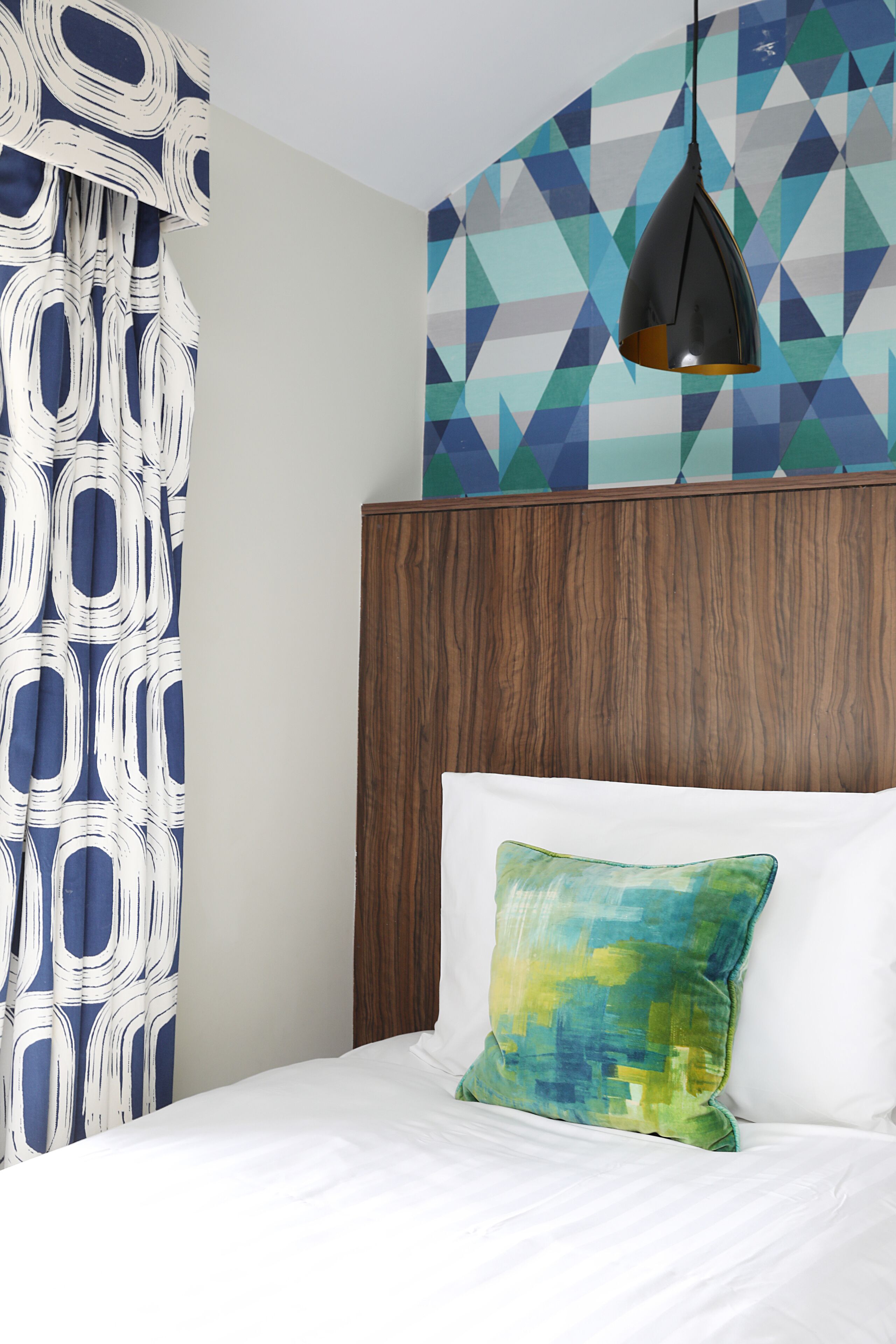 A bedroom with a wooden headboard, a white bed, a green and blue pillow, and a geometric-patterned wall.