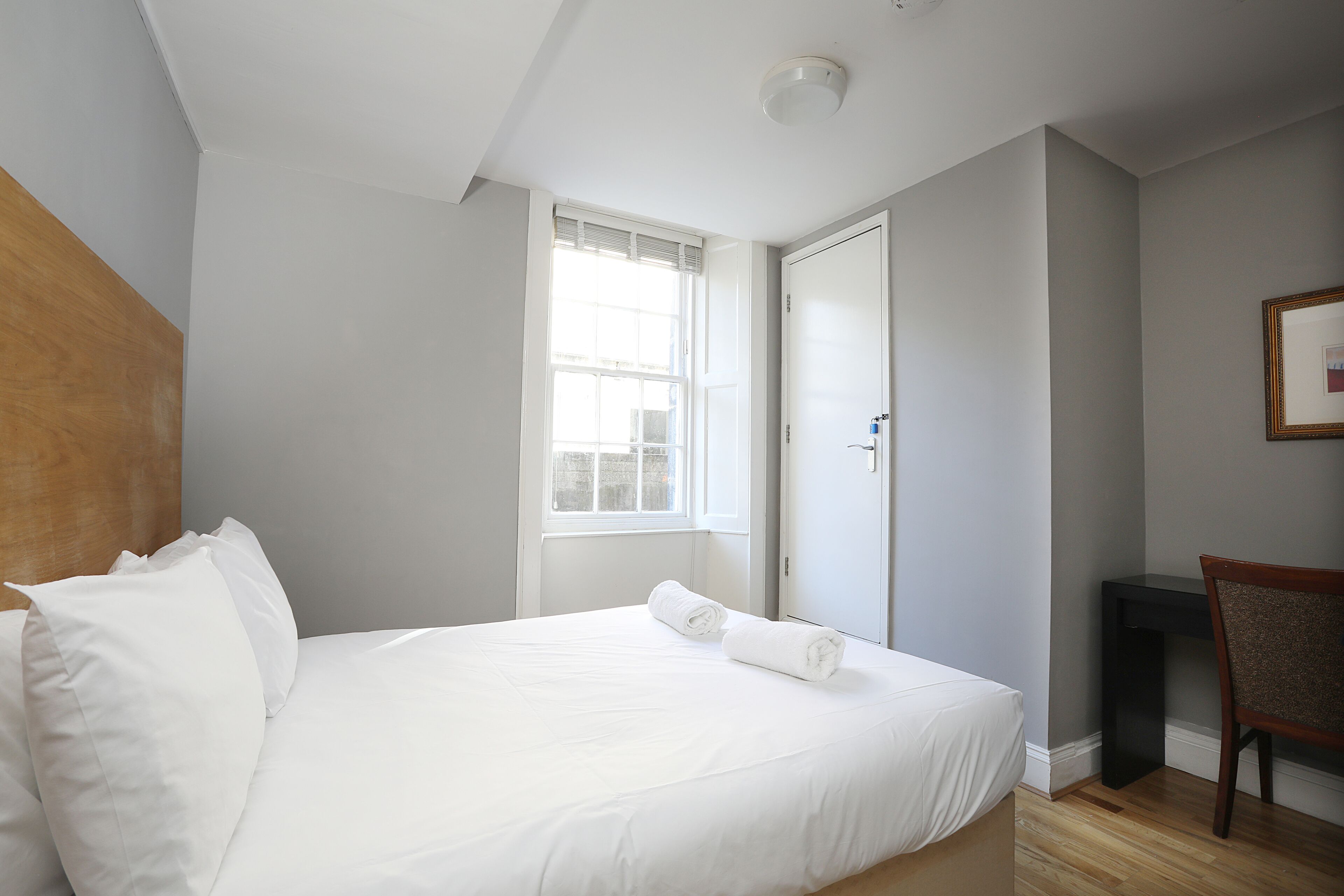 A neatly made bed with white linens, a wooden headboard, a window with blinds, a door, and a small desk with a chair.