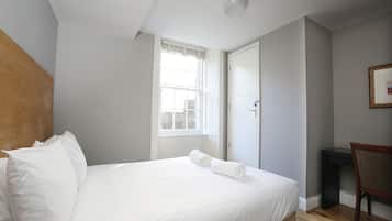 A neatly made bed with white linens, a wooden headboard, a window with blinds, a door, and a small desk with a chair.