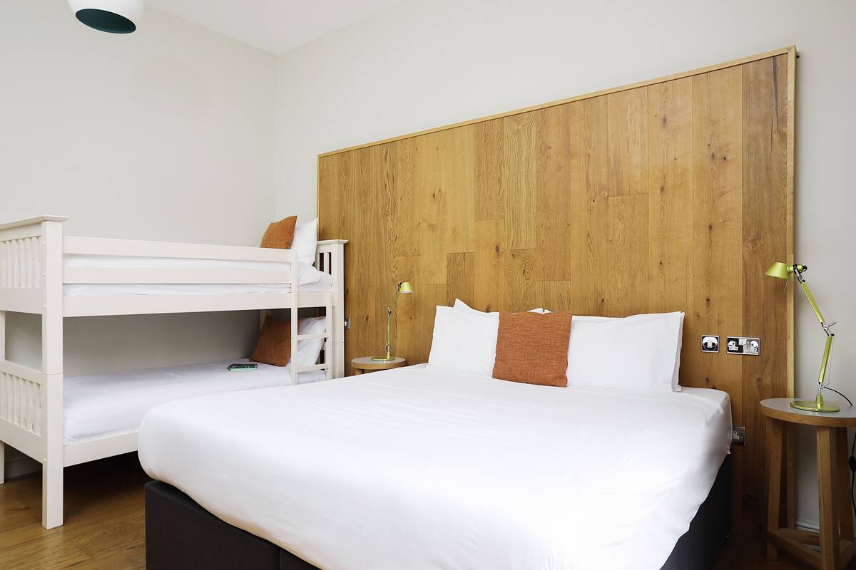 Ett hotellrum med en stor säng, en mindre våningssäng, två sängbord och en sänggavel i trä.