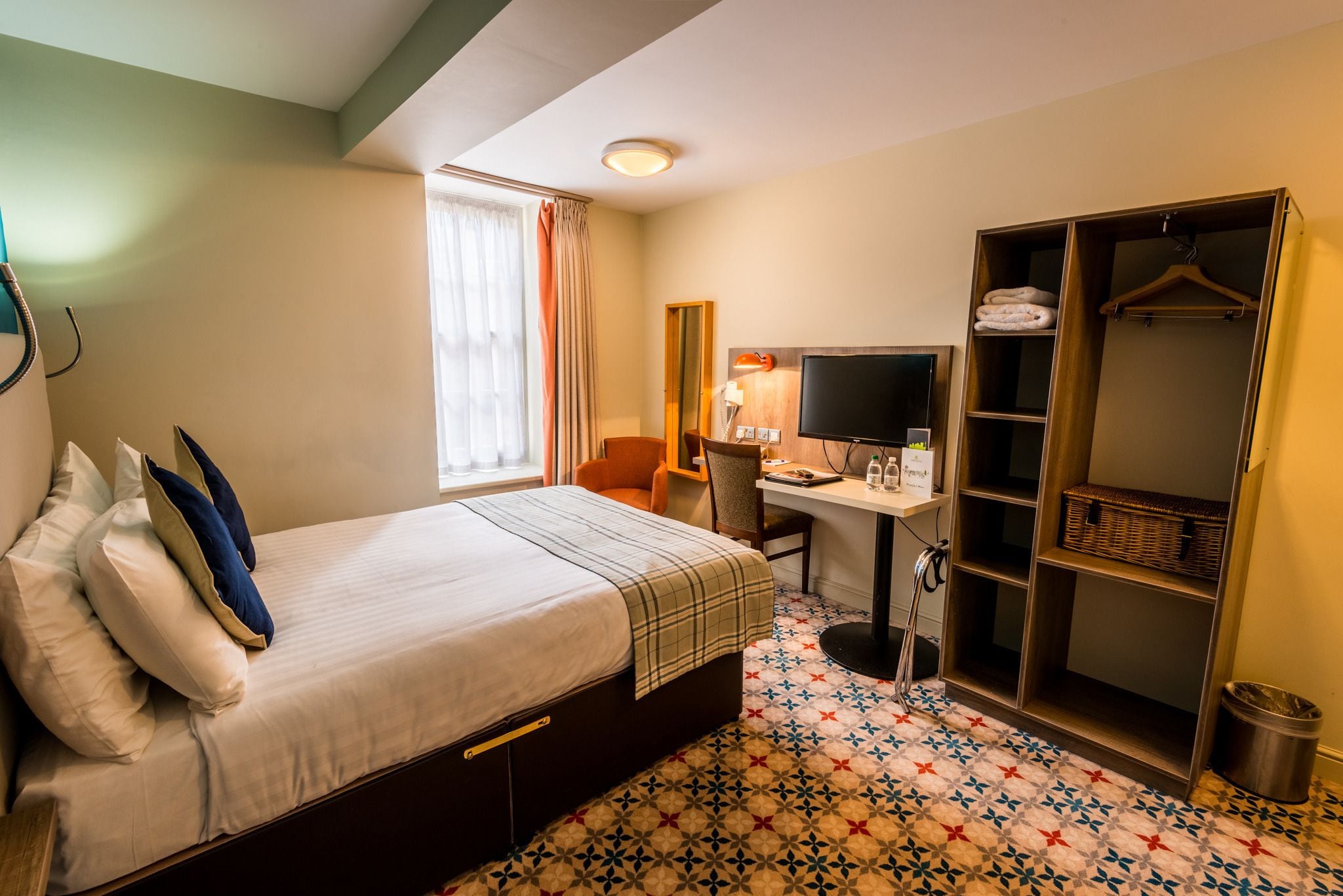 Ett hotellrum med en säng, ett skrivbord, en stol, en TV, en garderob och ett golv med ett mönster.