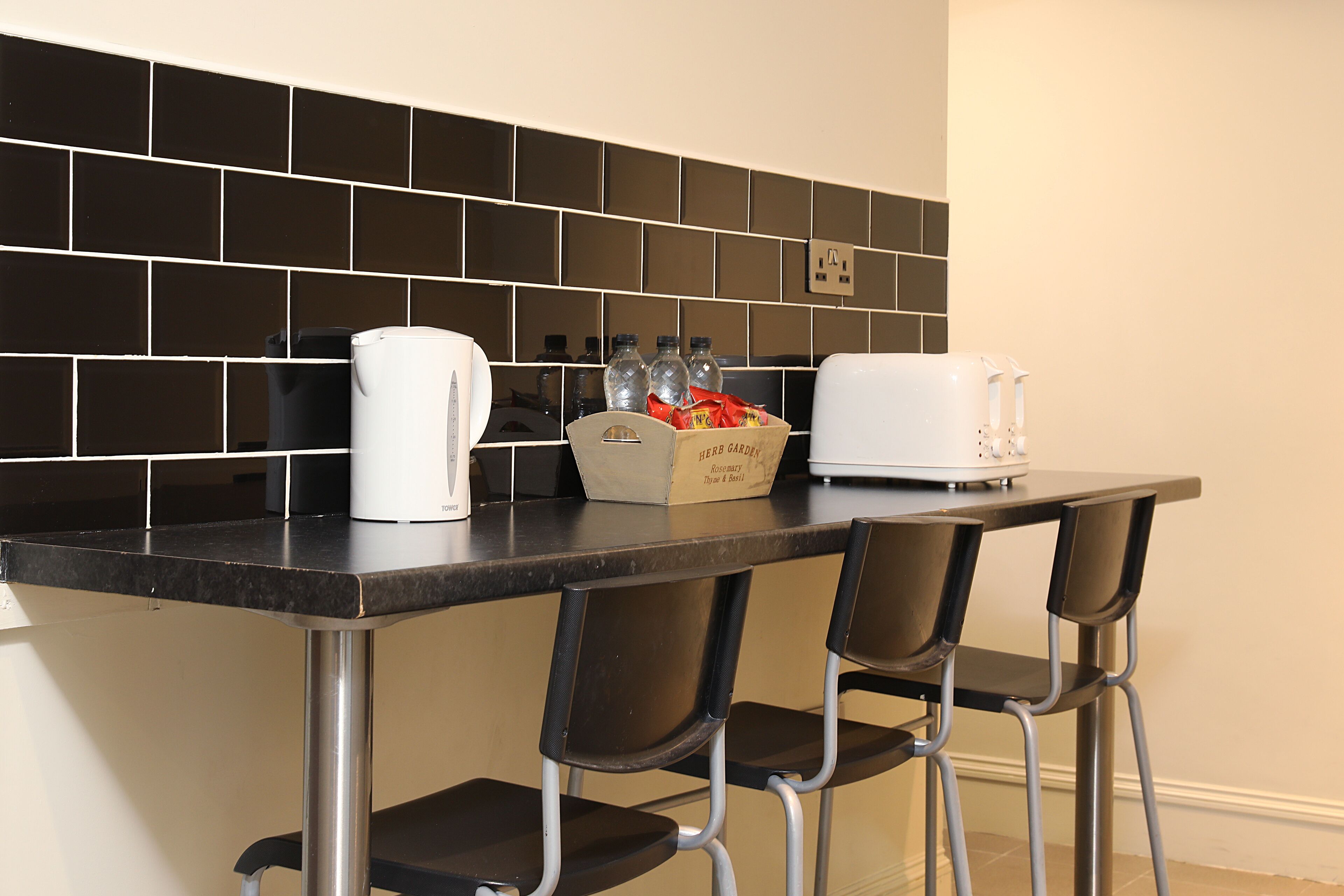 A kitchen with a black tiled backsplash, a white coffee maker, a toaster, and a box of bread. There are three black chairs at the counter.