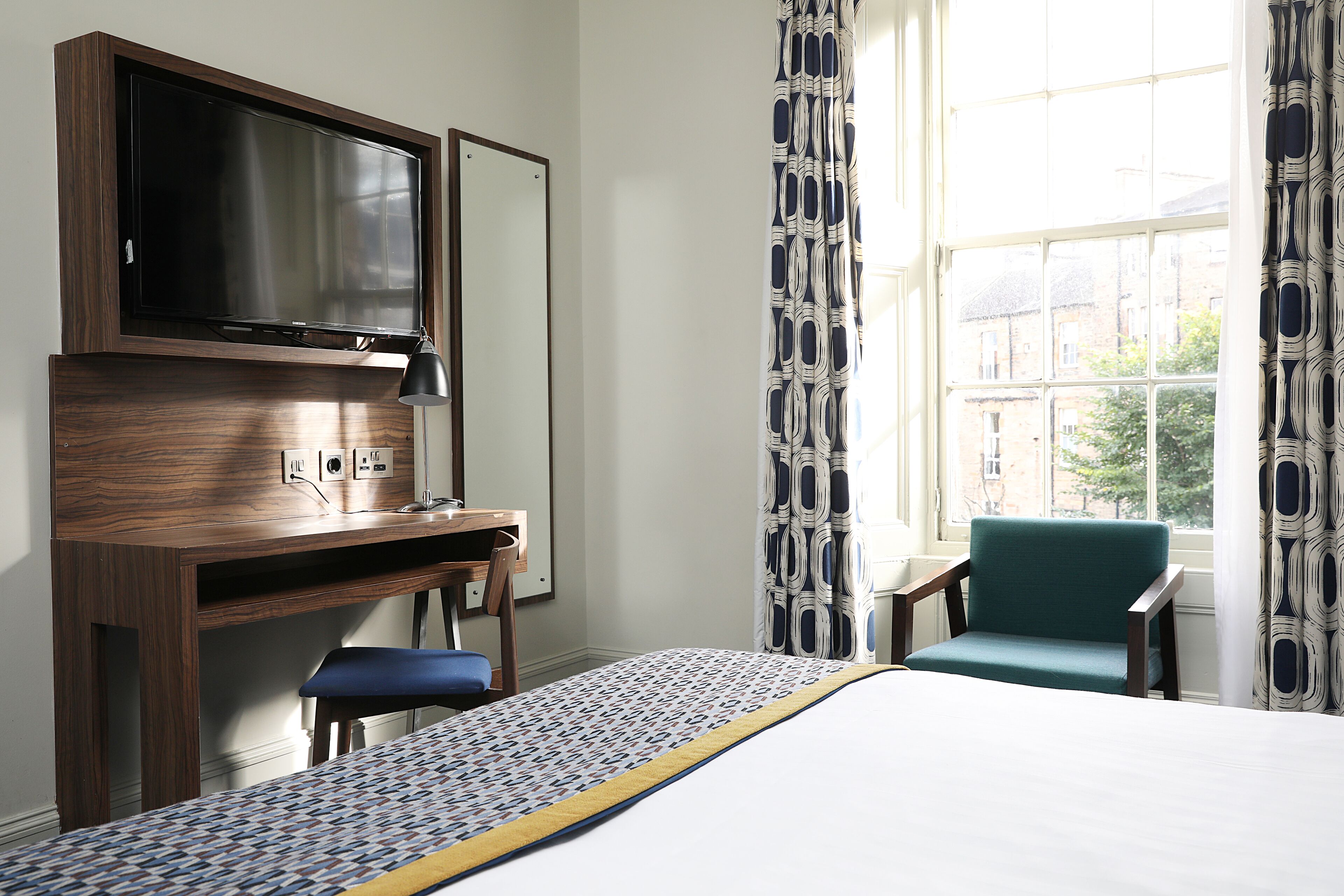 A hotel room with a bed, a desk with a TV, a chair, and a window with curtains.
