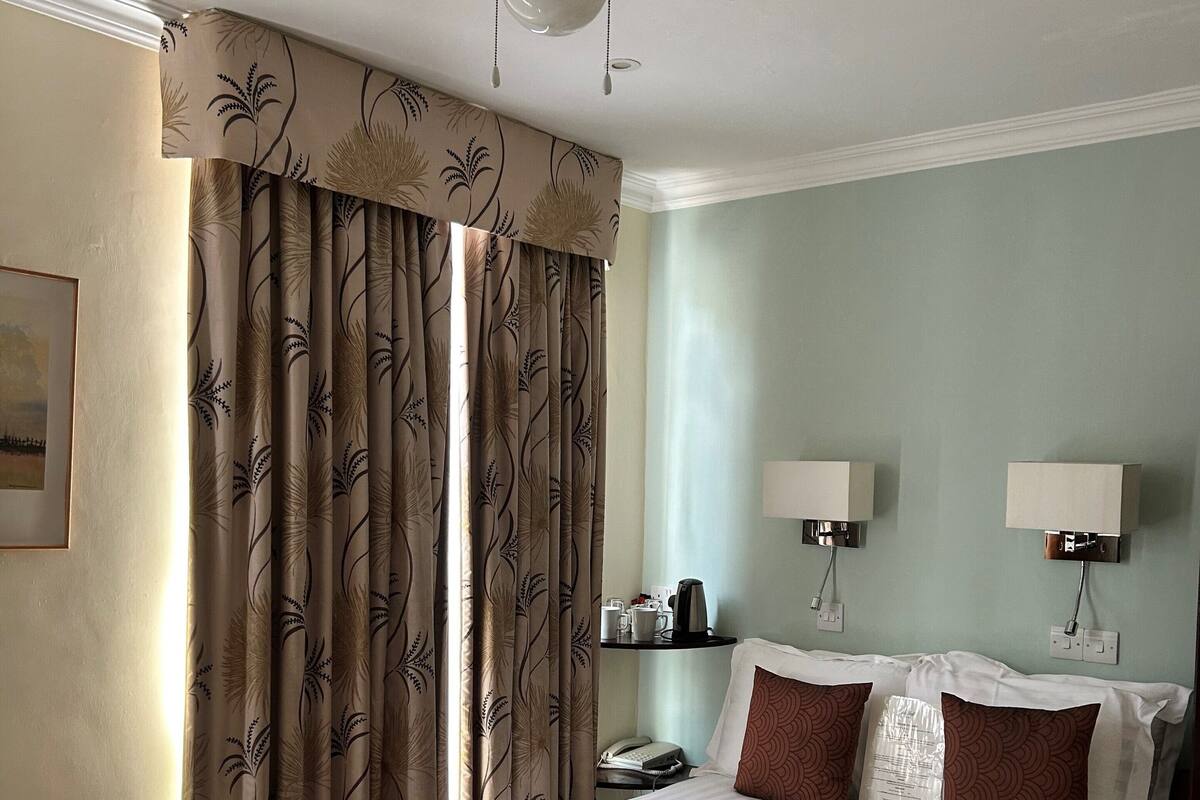 A hotel room with a bed, a ceiling fan, a nightstand with a lamp, and a window with curtains.