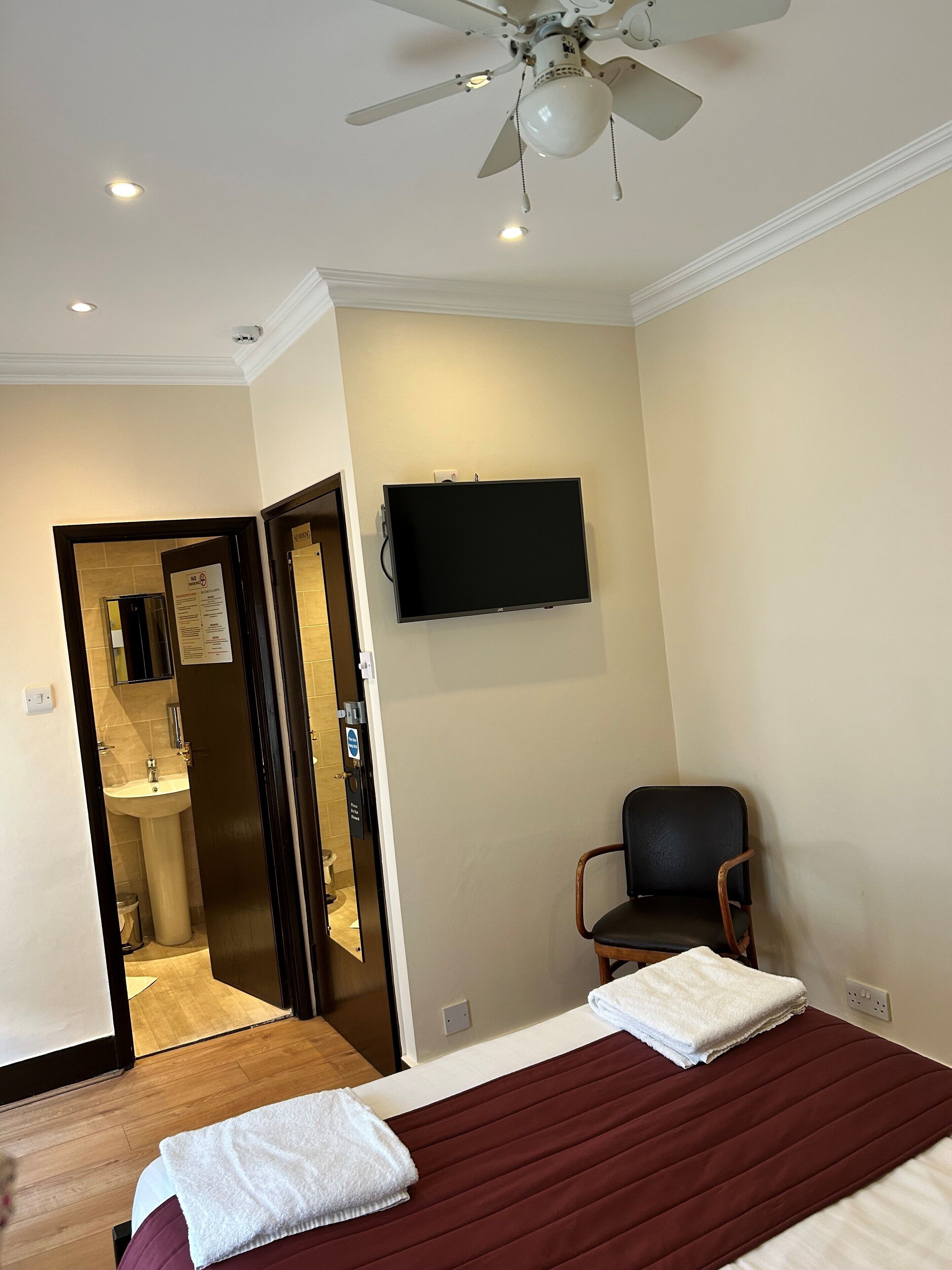 Ein Hotelzimmer mit einem Bett, einem Sessel, einem Fernseher, einem Deckenventilator und einem durch eine offene Tür einsehbaren Badezimmer.