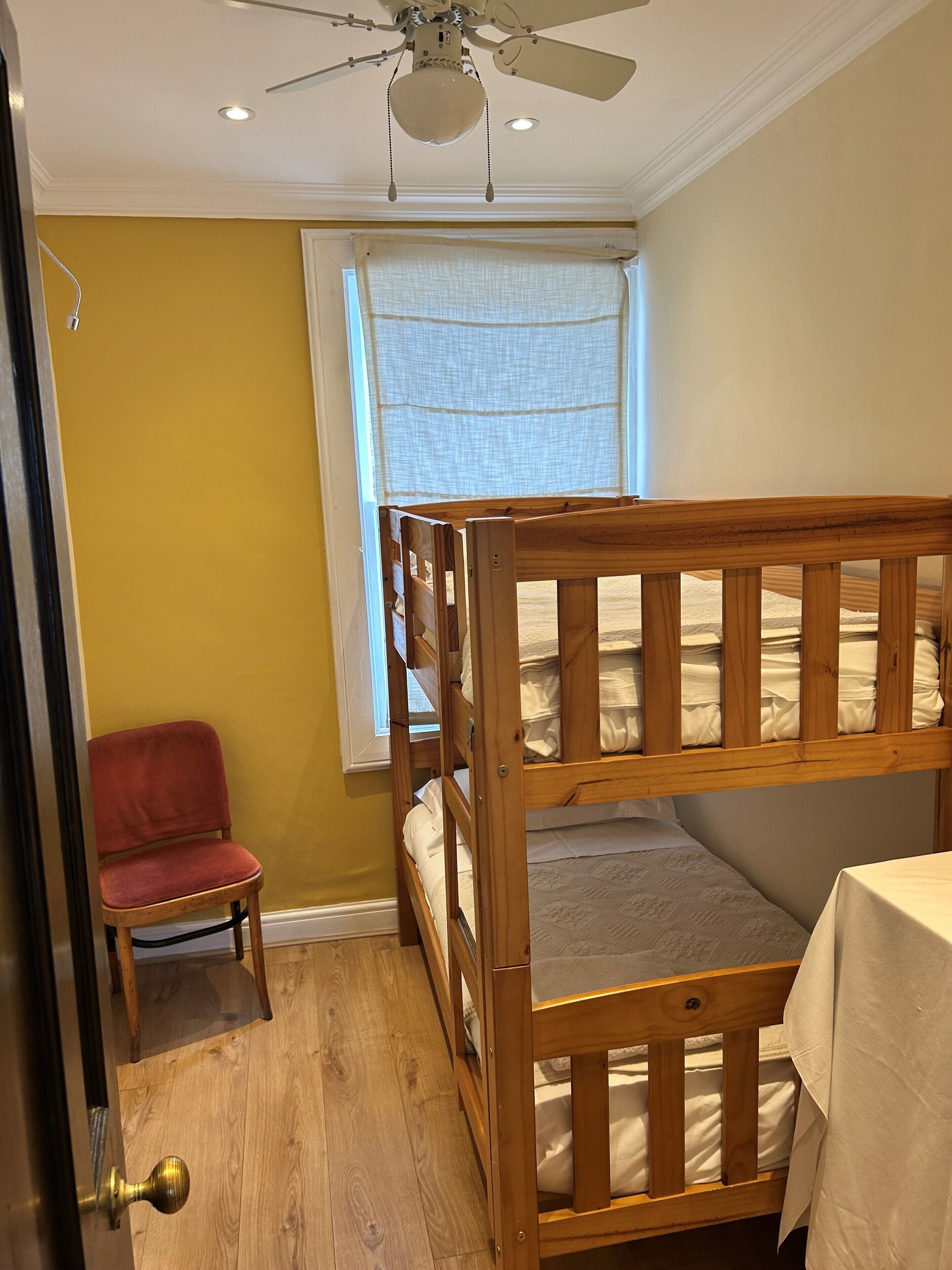 A room with a bunk bed, a chair, a table, a ceiling fan, and a window with a curtain.
