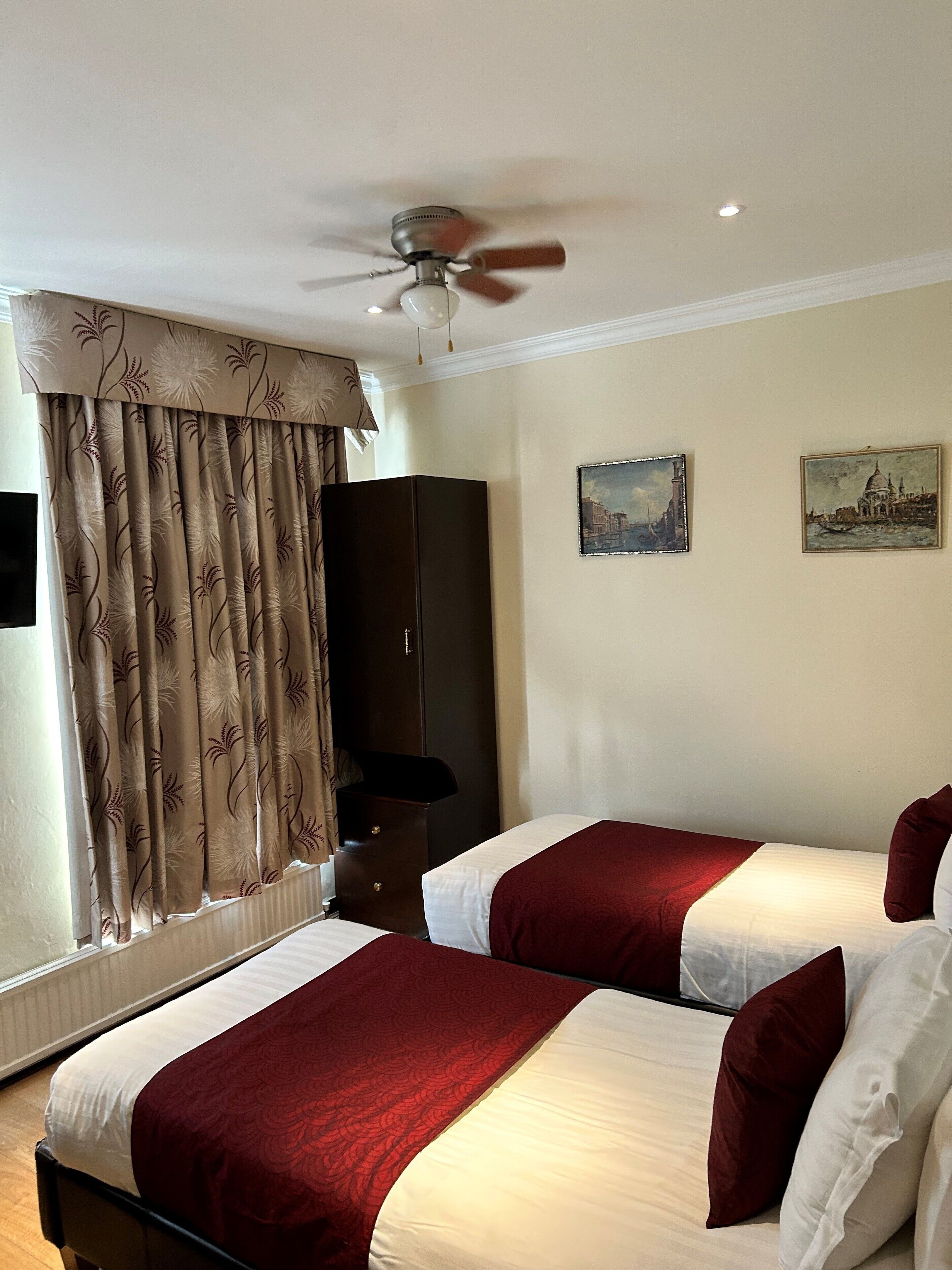 A hotel room with two beds, a ceiling fan, a large window with curtains, and two paintings on the wall.