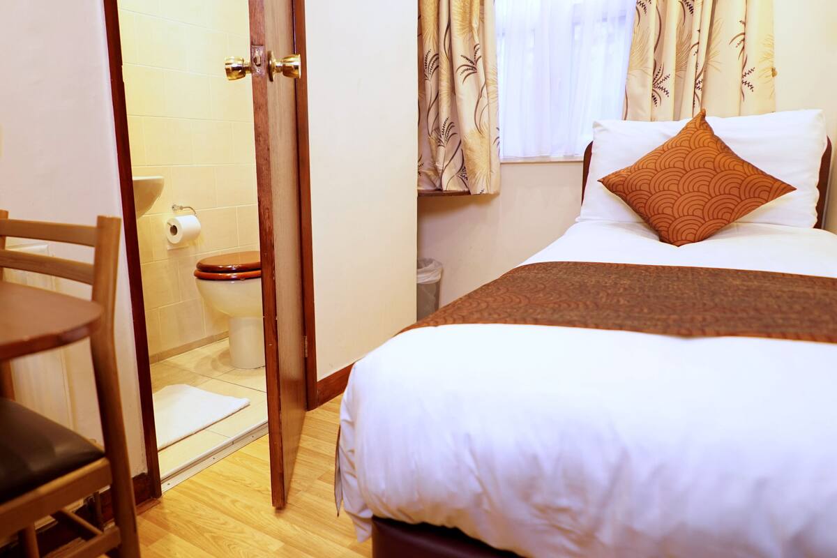 A hotel room with a bed, a wooden chair, a table, a toilet, and a shower area.