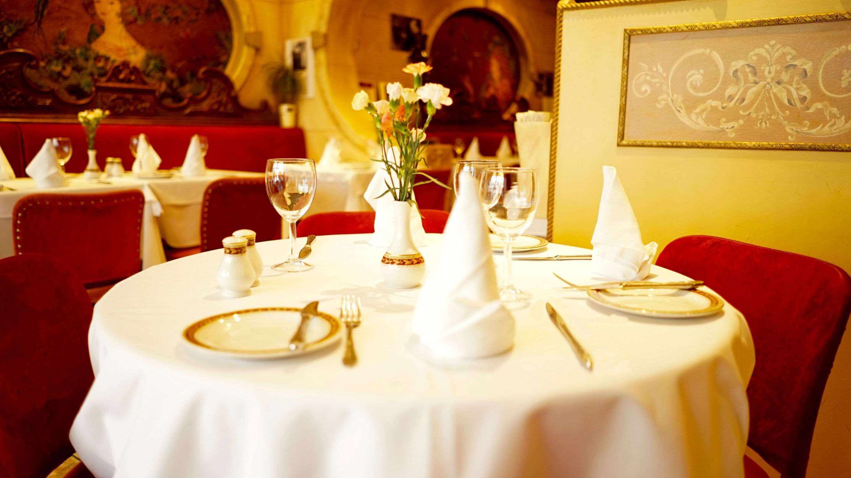 Mesa de comedor preparada para cuatro, con manteles blancos, platos, cubiertos y un florero con flores.