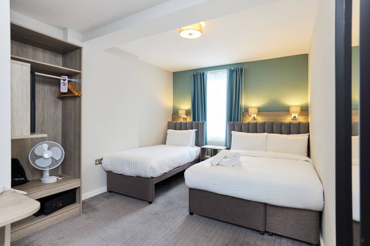 A hotel room with two beds, a desk, a fan, and a wardrobe.
