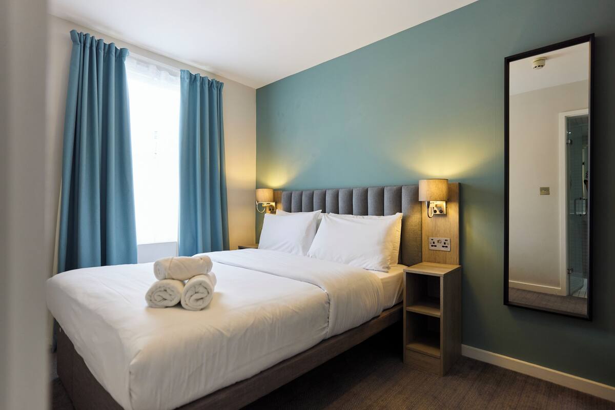A neatly made bed with white linens and rolled towels, a bedside table with a lamp, and a large mirror on the wall.
