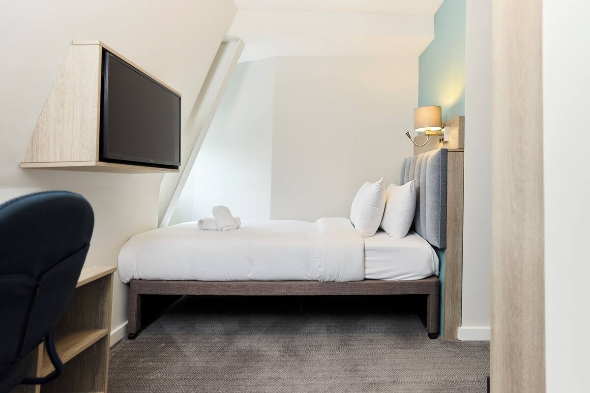 A modern hotel room with a bed, a desk, and a TV mounted on the wall.