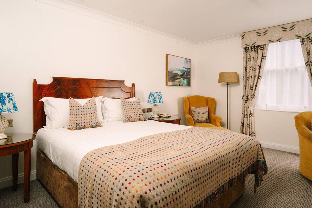 Habitación de hotel con una cama de madera, un sillón amarillo, una mesita de noche, una lámpara y un cuadro en la pared.
