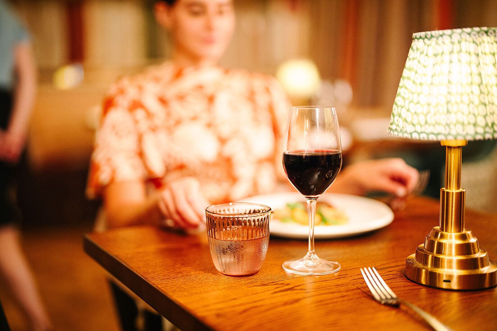 A dining table with a glass of red wine, a glass of water, and a table lamp.