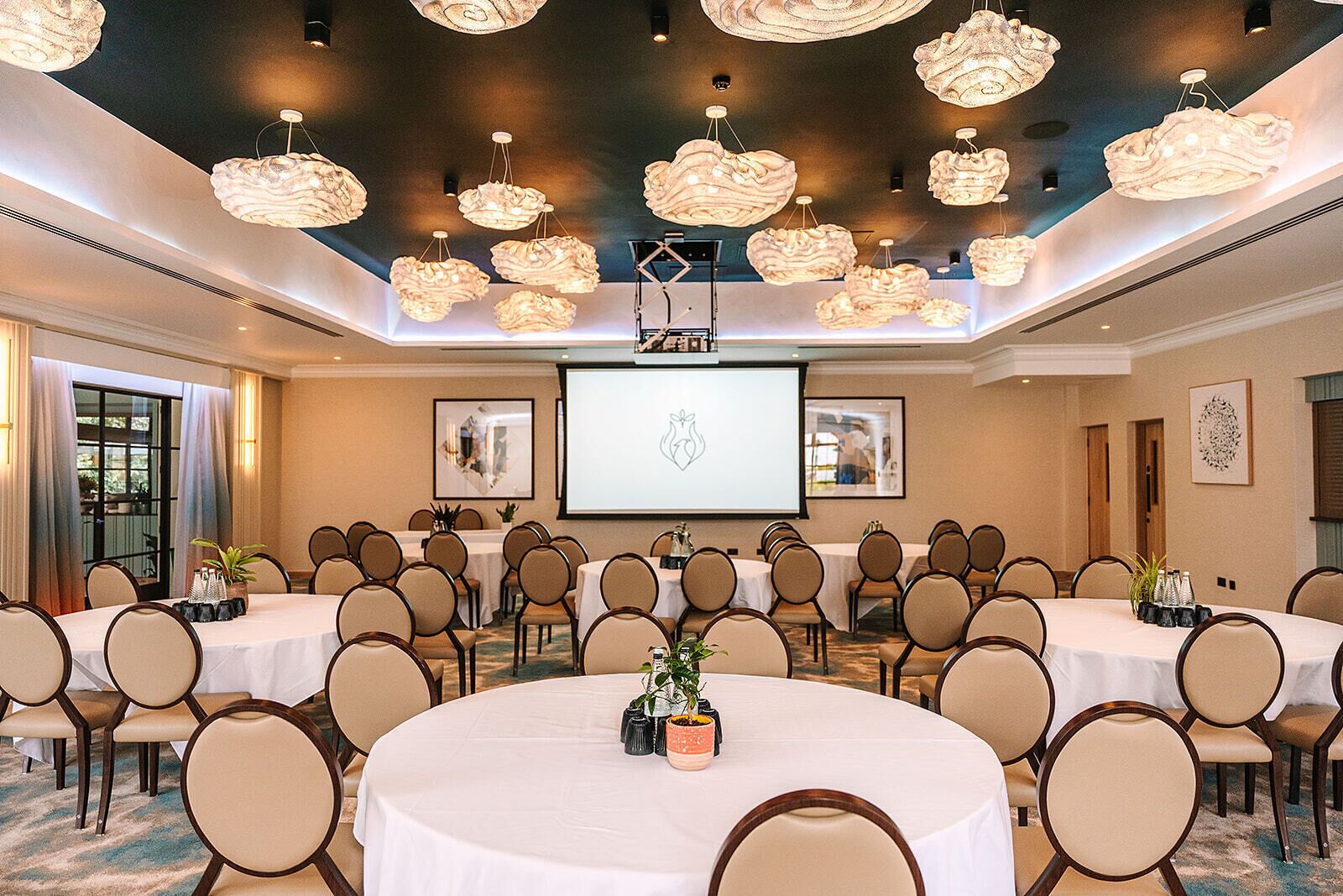 Een vergaderruimte met meerdere ronde tafels, beige stoelen en een groot scherm voor presentaties.