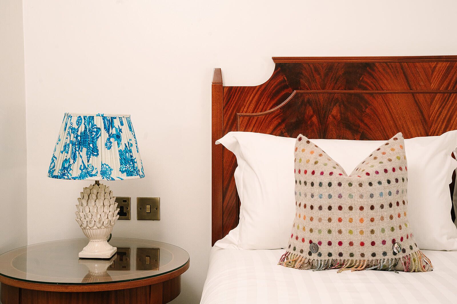 A hotel room with a wooden headboard, a round bedside table with a lamp, and a bed with white bedding and a colorful polka dot pillow.