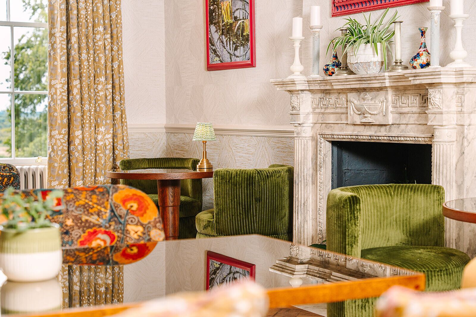 A room with a fireplace, green upholstered chairs, a table with a lamp, and a window with curtains.