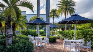 Un espace extérieur aménagé en terrasse, avec des tables et des chaises, entouré de palmiers et de haies.