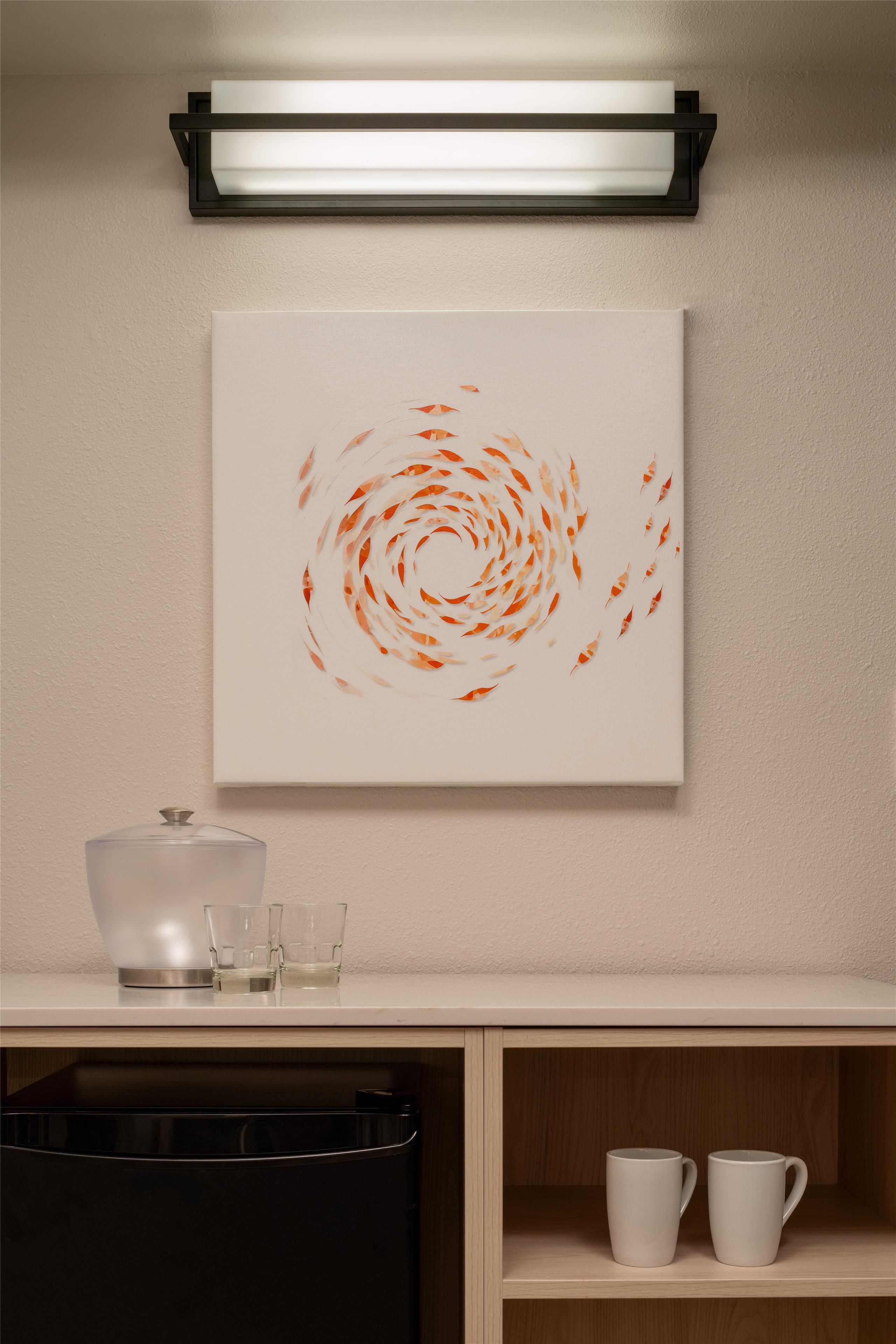 A wall-mounted artwork with a circular pattern, a kitchen counter with a kettle, glasses, and a cabinet below.
