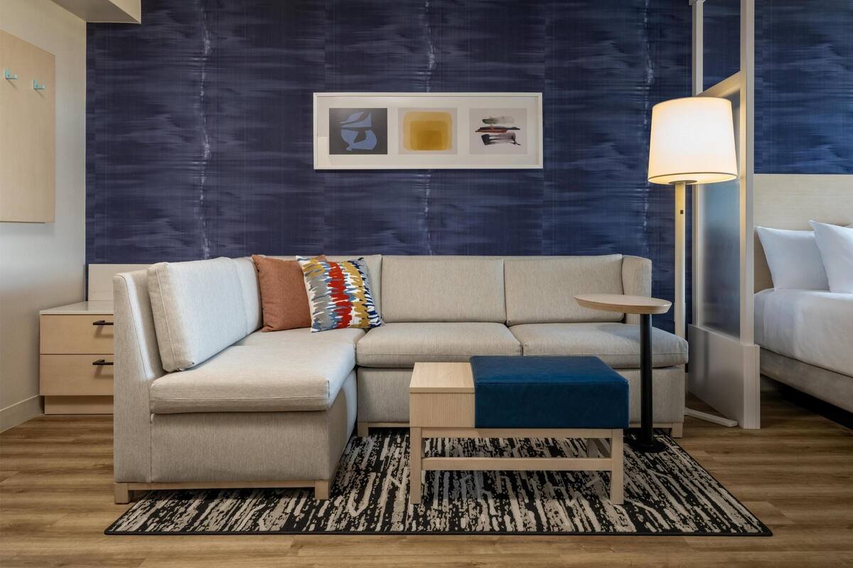 Een moderne woonkamer met een hoekbank, een klein bijzettafeltje, een vloerlamp en een lijstwerk op de muur.
