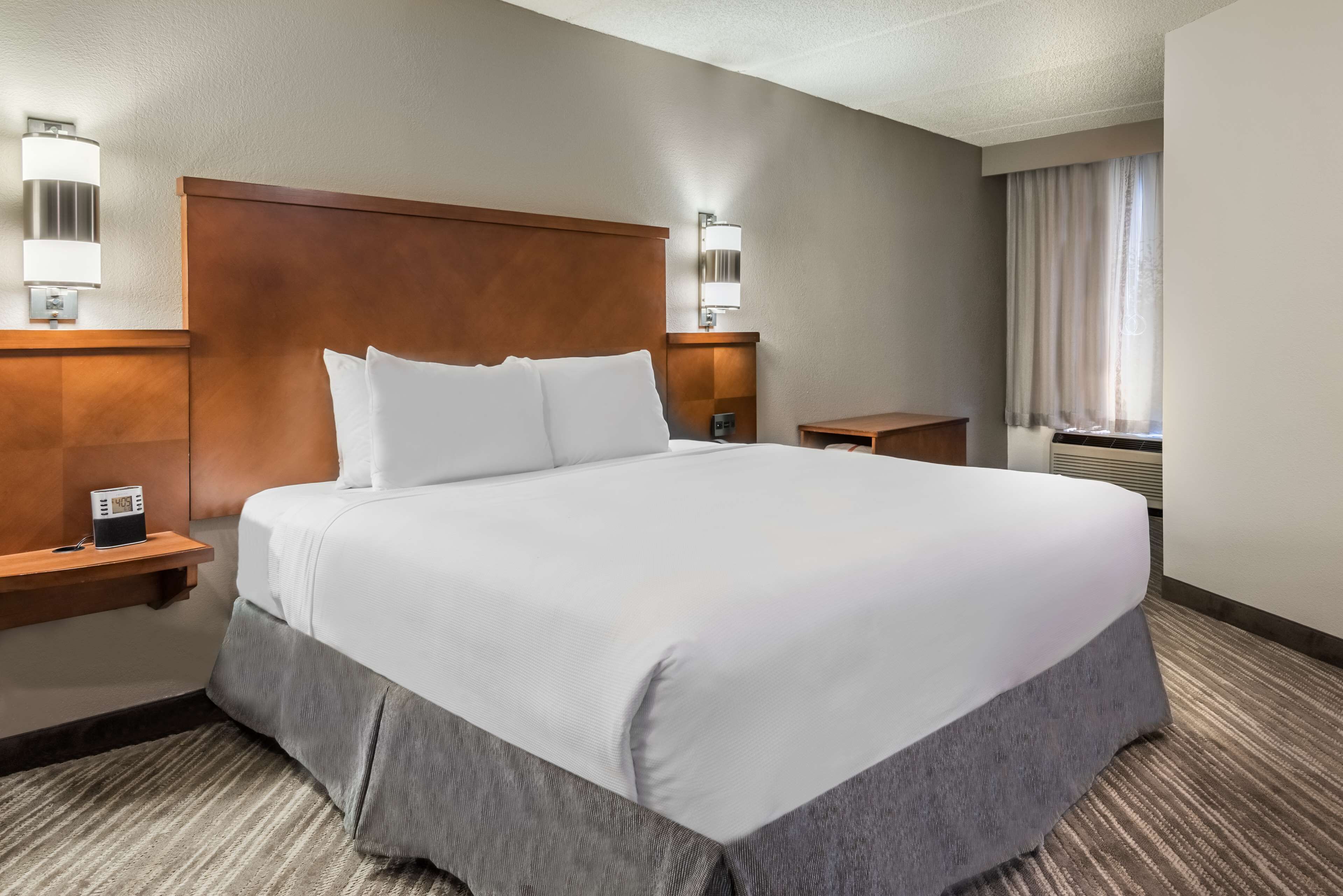 A hotel room with a large bed, two bedside tables, a nightstand, a wall-mounted lamp, and a window with curtains.