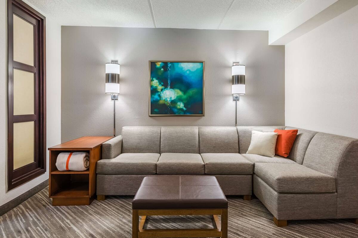 Une chambre d’hôtel moderne avec un canapé d’angle gris, une table d’appoint en bois, un banc et un grand tableau abstrait accroché au mur.