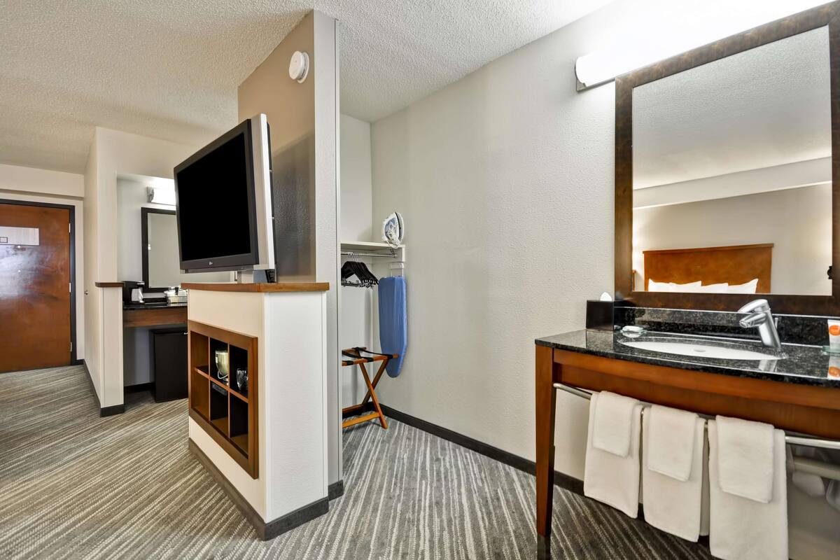 Une chambre d’hôtel équipée d’une télévision à écran plat, d’une petite kitchenette, d’une salle de bain avec un lavabo et un miroir, et d’une tête de lit en bois.