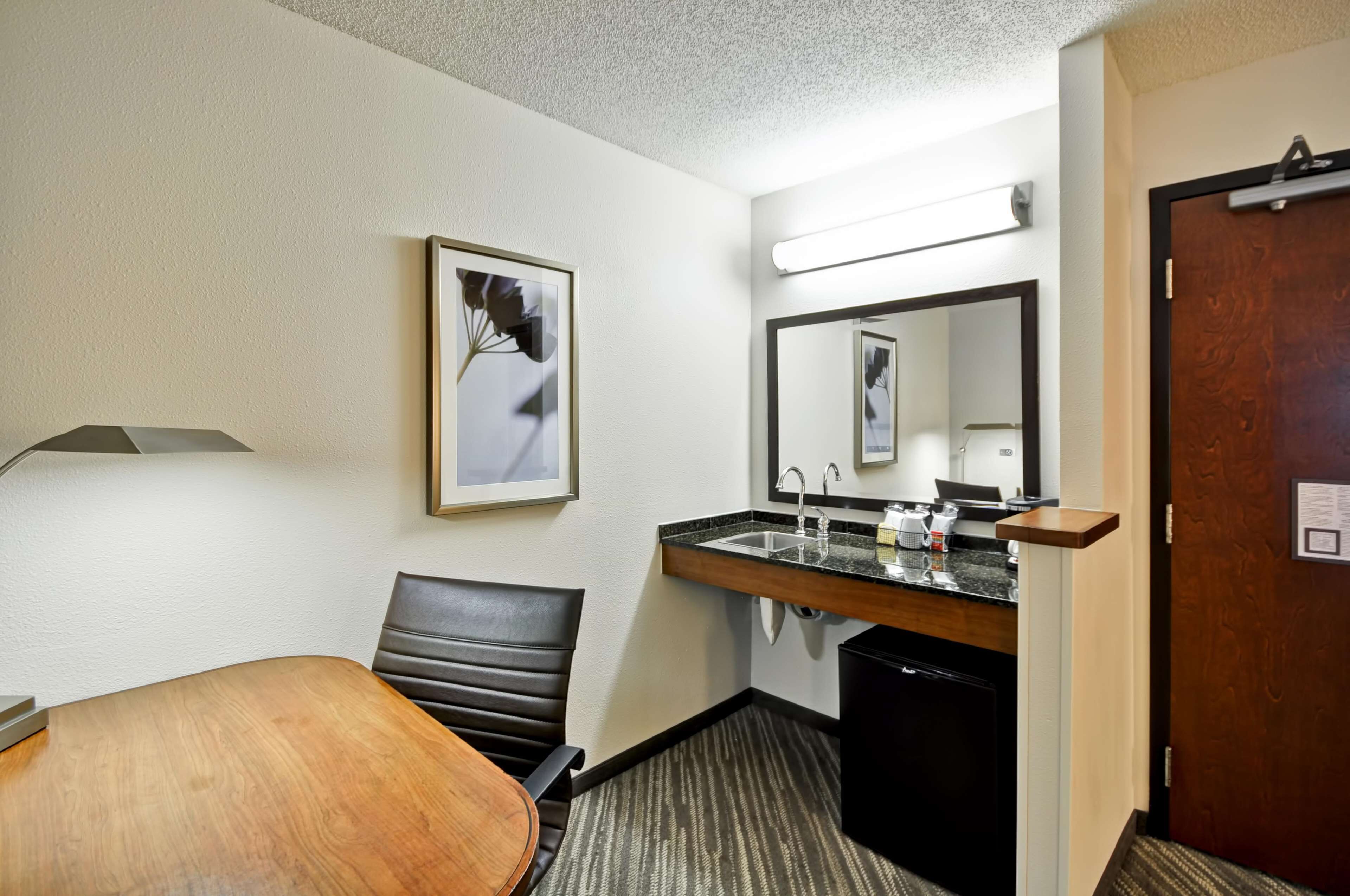 Una habitación de hotel con una mesa de comedor de madera, una silla de cuero negro, un lavamanos empotrado con espejo y una puerta con marco de madera.