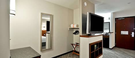 A hotel room with a flat-screen TV, a mirror, a small seating area, and a door to the right.
