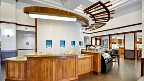 Un vestíbulo de hotel moderno con un mostrador de recepción de madera en el centro, una lámpara grande y una exhibición de alimentos.