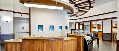 A modern hotel lobby with a central wooden reception desk, a large light fixture, and a food display.